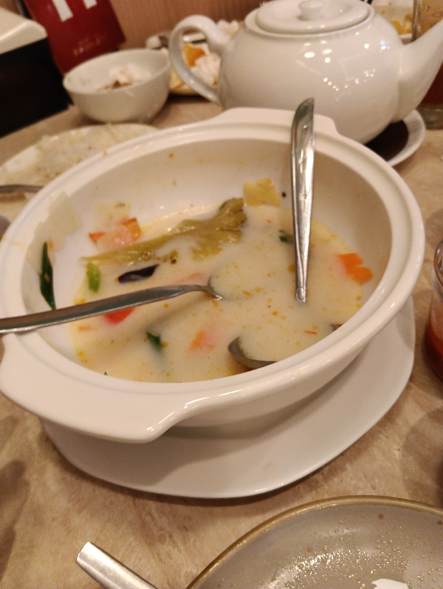 A close-up, slightly overhead shot captures a nearly empty white soup bowl containing a creamy broth, some scattered vegetable pieces, and two silver spoons, one resting in the bowl and the other partially submerged. The bowl is placed on a white saucer on a light brown, textured surface that appears to be a table. In the background, out of focus, is a white ceramic teapot with a handle and lid. A hint of a red object is visible in the far left background. The scene is indoors, suggesting a dining setting. The image does not provide any information to place it specifically in Sungai Raya, Indonesia.