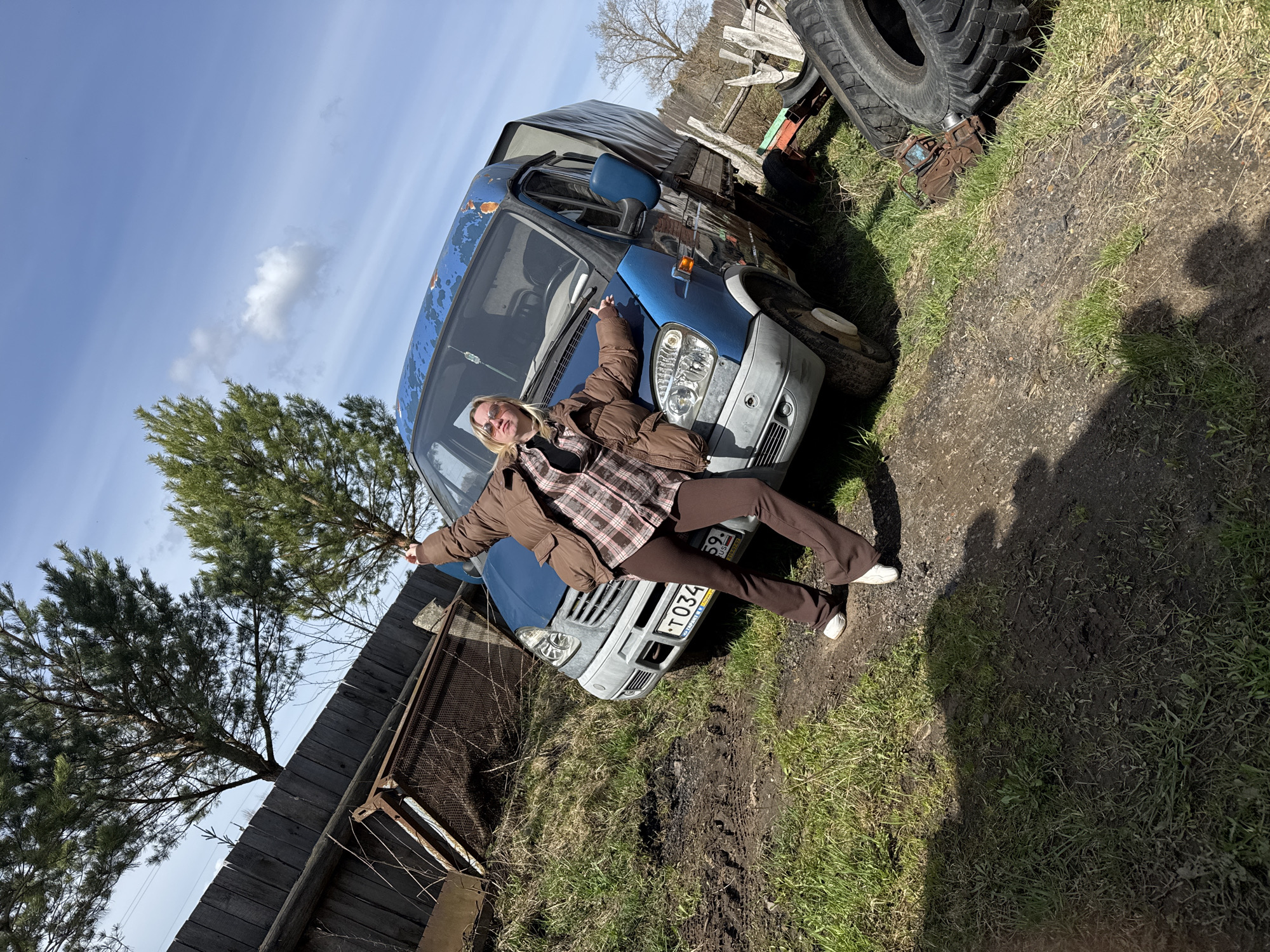 A person is playfully posing on the front of an aged, light blue cargo van, captured outdoors under a bright blue sky. The individual, appearing to be a young adult with blonde hair and sunglasses, wears a brown puffer jacket over a plaid shirt and brown pants with white sneakers. They lean against the hood and windshield of the van, with arms outstretched and one leg extended, conveying a relaxed or whimsical mood.
The van itself shows signs of wear, with peeling paint on its blue body and a grey bumper. It is parked on an unpaved, uneven ground surface, characterized by patches of sparse green grass, dirt, and some muddy areas with visible tire tracks.
In the background, a rustic dark wooden plank fence runs along the left side. Several evergreen trees (pines) and some bare deciduous trees are visible against the clear sky, which features a few small, wispy white clouds. To the right of the van, large dark tires are stacked on the ground, alongside some rusty metal objects. Bright sunlight illuminates the scene, casting distinct shadows, indicating a sunny day, likely midday or early afternoon.
A Russian license plate is visible on the front of the van, displaying "T 034 59 RUS", with "RUSSIA" printed below the numbers. The setting, with its unpaved ground, rustic fence, and older vehicle, suggests a rural or semi-rural location.
