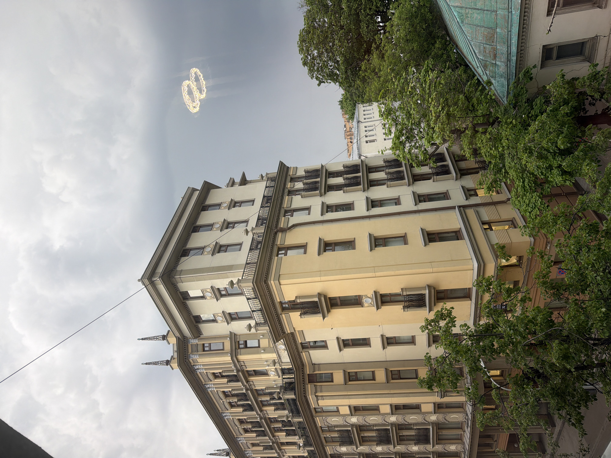 The image captures a building in Moscow, Russia, with a cloudy sky as the backdrop. In the sky, a pair of glowing, interconnected rings, resembling wedding rings, float above the structure. The architecture of the building is intricate with a multi-tiered design, incorporating both light yellow and beige shades. Windows and small balconies adorn the façade, adding depth. Verdant trees are positioned to the right, framing the building, and a glimpse of a roof is visible. The image is taken from a low angle, emphasizing the height of the building.