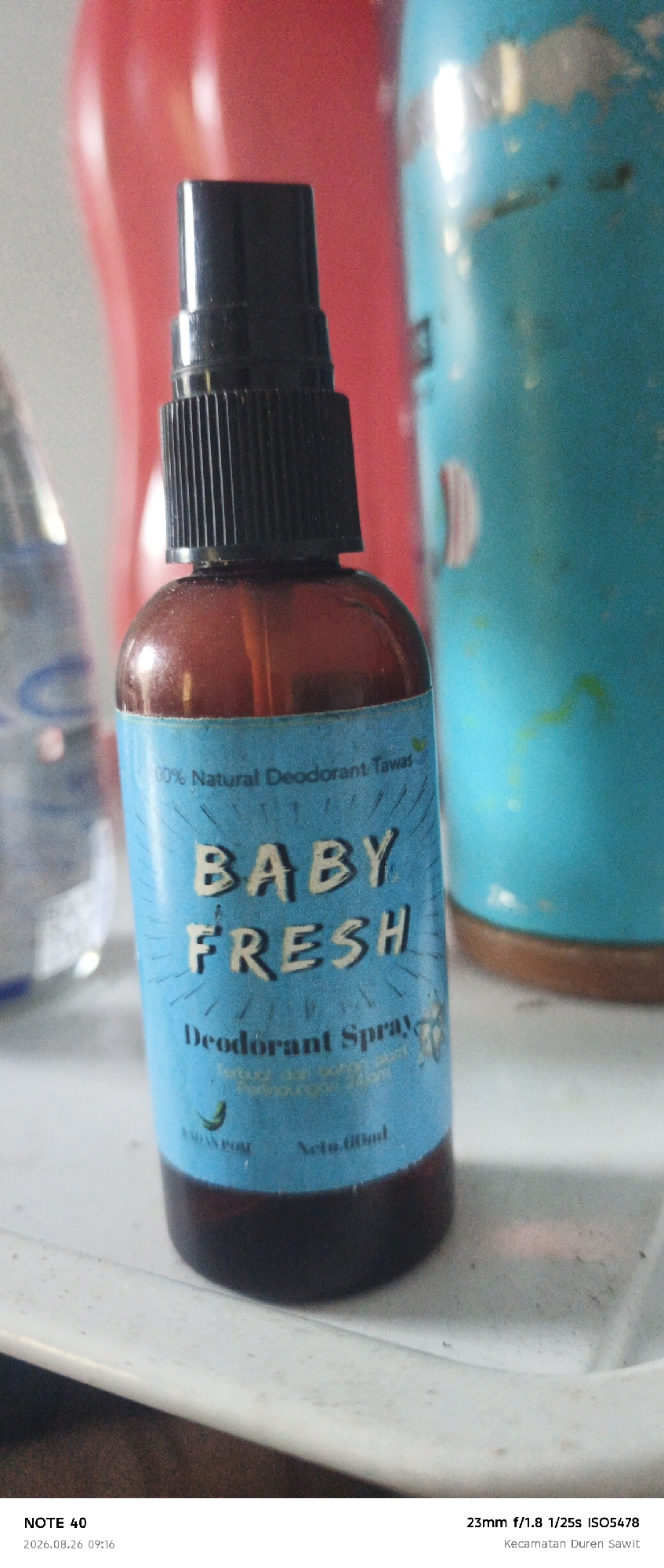 A close-up, eye-level shot features a spray bottle of "BABY FRESH Deodorant Spray" in the foreground. The bottle has a blue label with white text and is partially filled with a brown liquid. The bottle is positioned on a white surface, possibly a tray or shelf, which shows signs of dust and wear. In the background, out of focus, are colorful objects, including a bright red curved surface and a teal-colored cylindrical object with some lighter colored marks. The lighting appears to be artificial, casting highlights on the bottle and background elements. The bottom of the image contains metadata: "NOTE 40", a date and time "2026.08.26 09:16", camera settings "23mm f/1.8 1/25s ISO5478", and a location "Kecamatan Duren Sawit".