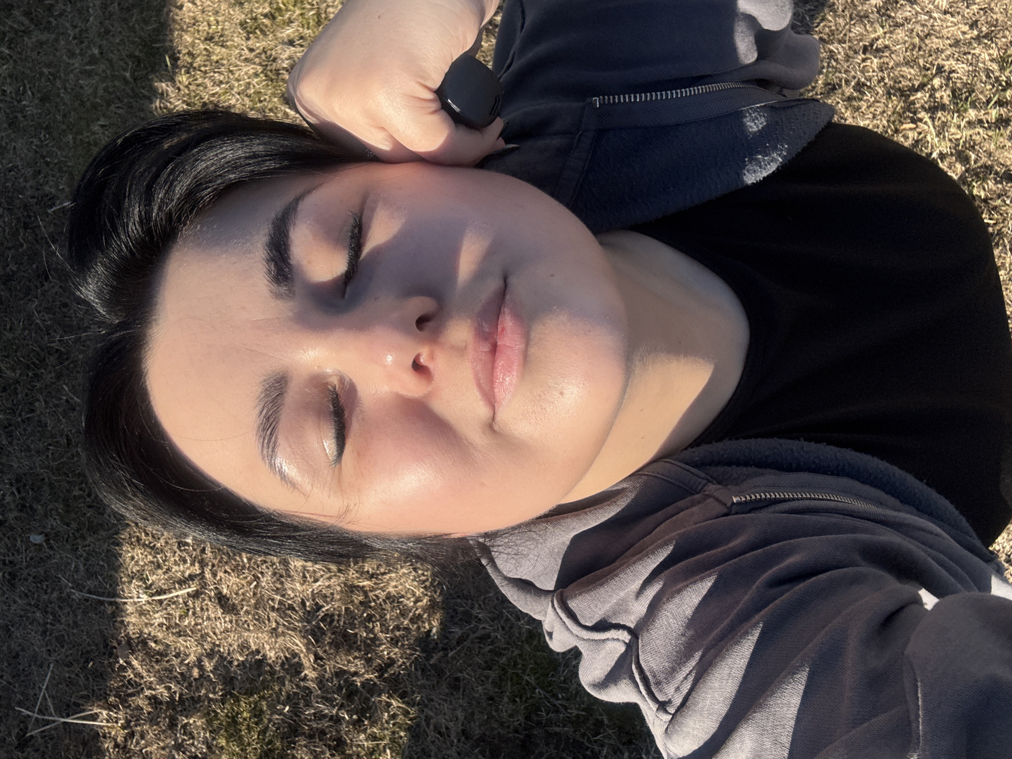 An overhead shot shows a person, likely female, lying peacefully on dry, sparse grass in Chaikovsky, Russia. The individual has dark hair and is wearing a black t-shirt beneath a grey, zip-up hoodie. Their head is tilted slightly back, and their eyes are gently closed, suggesting relaxation or enjoyment of the sun's warmth. One hand is partially visible near the head, holding a small, black object, possibly an earbud or a device.
The scene is illuminated by strong, direct sunlight, indicating a clear, sunny day, likely during mid-morning or afternoon. This harsh light creates distinct shadows on the grass and highlights the person's face. The dry, patchy grass suggests an outdoor setting like a park or field, possibly in a season with less lush vegetation. The overall emotion conveyed is one of tranquility and serenity. No visible text is present in the image.