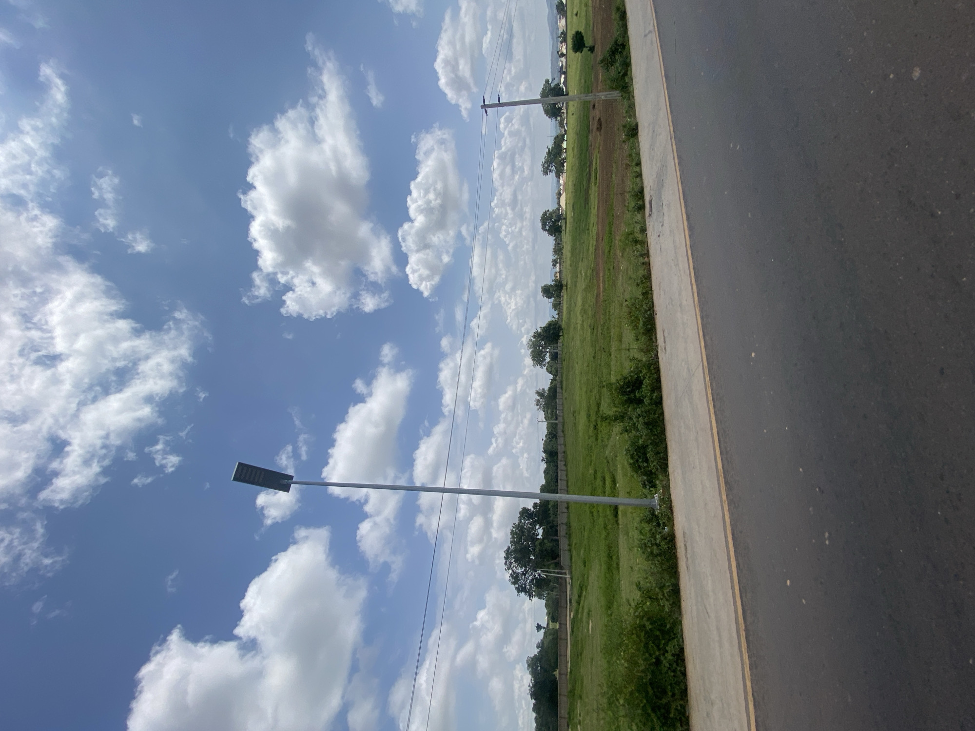 The image depicts a clear daytime scene with a bright blue sky dotted with fluffy white clouds. In the foreground, a paved road with a yellow line marking its edge is visible, sloping slightly upwards from left to right. Adjacent to the road is a concrete walkway. Beyond the walkway, a vast expanse of green grass stretches into the middle ground, with a few scattered trees and some low-lying bushes. A tall, slender lamppost stands prominently in the middle ground, positioned to the left of center. In the background, a line of trees and some buildings can be seen, suggesting a rural or semi-urban environment. The overall impression is one of an open, sunny landscape. The location is identified as Uke, Nigeria.