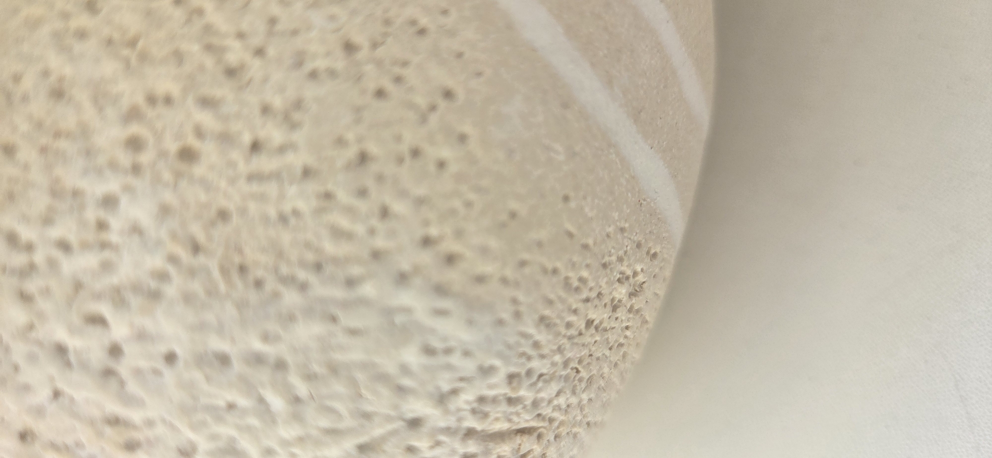 This close-up, macro-style image showcases a highly textured, light beige or tan surface. The surface is densely covered with numerous small, irregular indentations and pores, giving it a distinct bubbly or spongy appearance. This textured object dominates the left and central portions of the frame, curving diagonally towards the upper right.
On the right side of the image, the textured surface transitions to a smoother, lighter, off-white area, which appears to be either a background surface or another part of the same object. Two distinct, slightly raised, curved white lines or streaks are visible on the textured surface, running parallel to each other where it meets the smoother off-white section. These lines follow the object's curvature.
The overall impression is that of a detailed view of a porous material, strongly resembling a type of food known for its distinctive texture, such as injera (Ethiopian or Eritrean flatbread), which is characterized by similar "eyes" or holes. There are no people, explicit activities, time of day cues, weather details, or visible text in the image. The lighting is soft and even, highlighting the surface's intricate details.