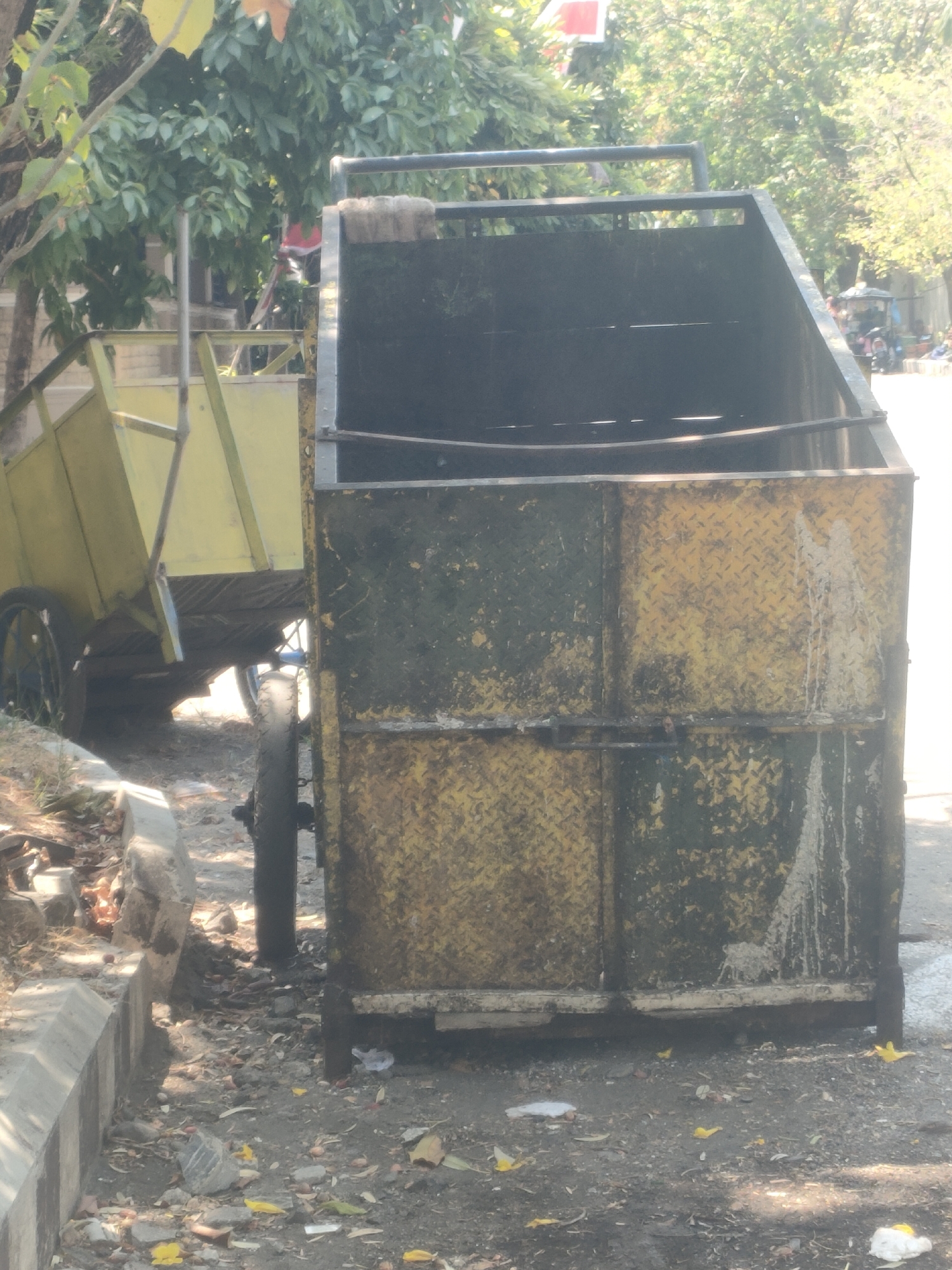 An open-top, rectangular metal dumpster, likely used for waste collection, stands in the foreground. It is predominantly yellow, with significant rust and patches of dark green or black grime covering its surfaces, particularly on the front panels where diamond plate texture is visible. The dumpster is positioned next to a curb and on a dirt and debris-strewn surface, suggesting an outdoor urban environment. To the left of the dumpster, a portion of a yellow, two-wheeled cart is visible, also appearing to be for waste transport, with a metal frame structure. The cart has at least one black wheel with spokes. Behind the dumpster, foliage from trees is apparent, with some sunlight filtering through, indicating an outdoor setting. In the distance, a street scene is faintly visible with hints of vehicles and possibly people, consistent with a city location. The overall impression is one of a utilitarian, weathered waste management area in an urban setting in Surabaya, Indonesia.