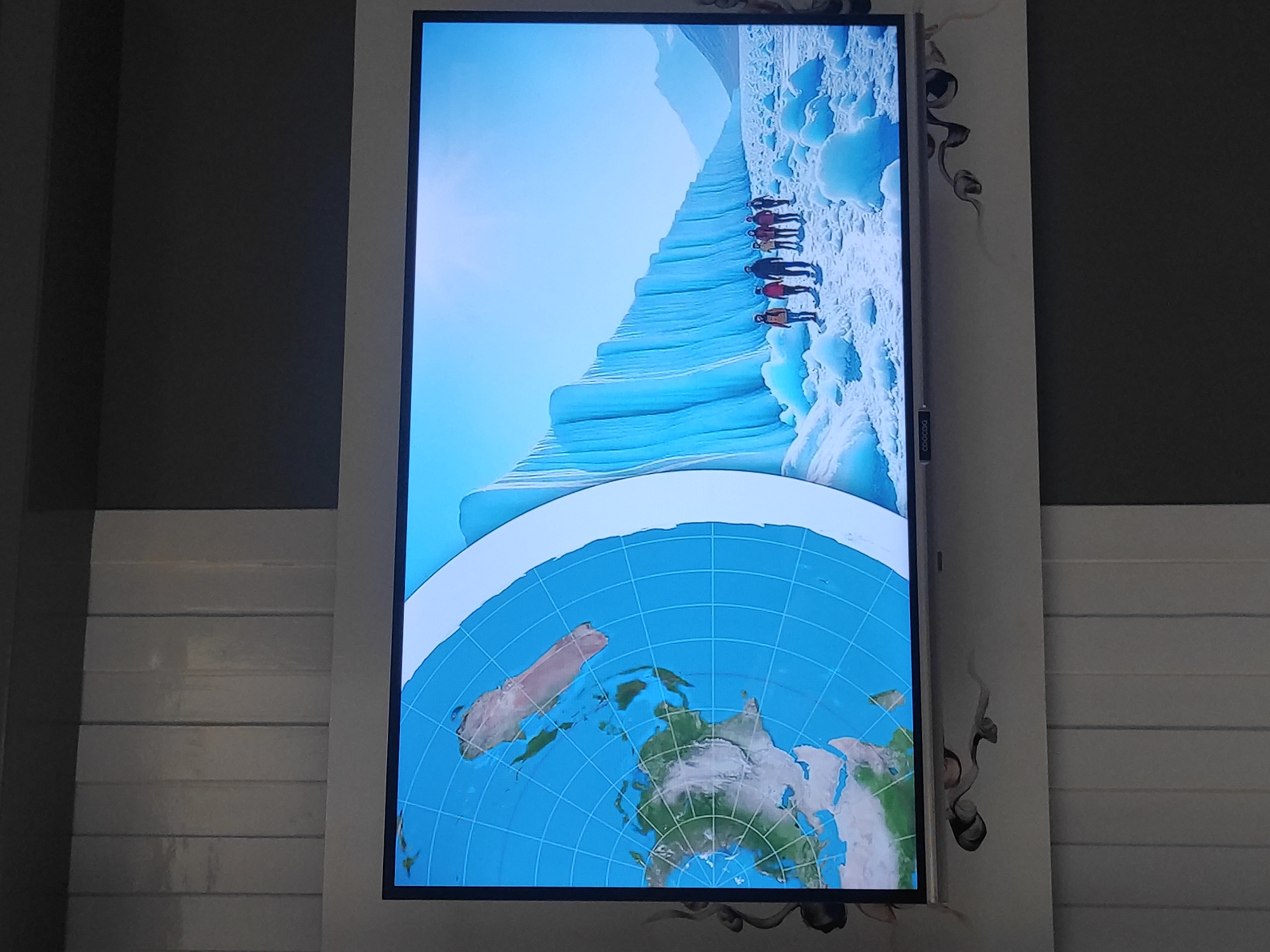 A large television screen displays a flat Earth map projected onto a blue grid. To the right, a group of seven individuals, dressed in outdoor clothing, walk along a snow-covered path. They are dwarfed by a towering, blue ice wall that stretches into the distance. The environment appears to be a cold, icy landscape with mountains visible in the background under a clear blue sky. There is no identifiable information to indicate this scene is located in Sumedang, Indonesia.