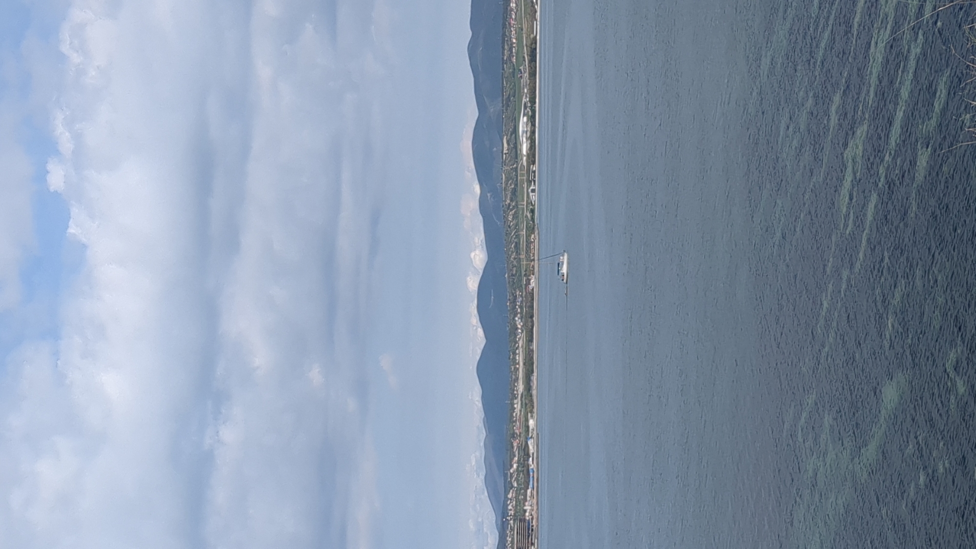 The image presents a serene panoramic landscape featuring a large body of water, a distant coastline backed by mountains, and a partly cloudy sky, captured during the daytime. The scene appears to be taken from an elevated viewpoint looking across a bay or sea.
In the upper section, the sky is a soft light blue, dotted with scattered white clouds. Some clouds are light and wispy, while others are larger and more defined, suggesting pleasant, clear weather.
Across the middle of the frame, a long horizon line reveals a distant landmass. This includes a developed coastline with visible buildings and structures, indicating a town or city, consistent with Gelendzhik, Russia. Behind this coastal strip, a range of dark mountains rises, their distant peaks appearing slightly hazy or lighter, blending into the atmosphere.
The majority of the lower and right portions of the image are dominated by the expanse of water, displaying a deep blue-green hue. On this calm surface, a white sailboat with a mast is visible, positioned somewhat centrally, suggesting it is gently sailing across the bay. Closer to the foreground (the right edge of the image), the water's surface shows textured patterns, possibly due to light reflecting off small ripples or submerged vegetation in shallower areas.
The overall impression is one of tranquility and openness, under bright, clear conditions. There is no visible text in the image.