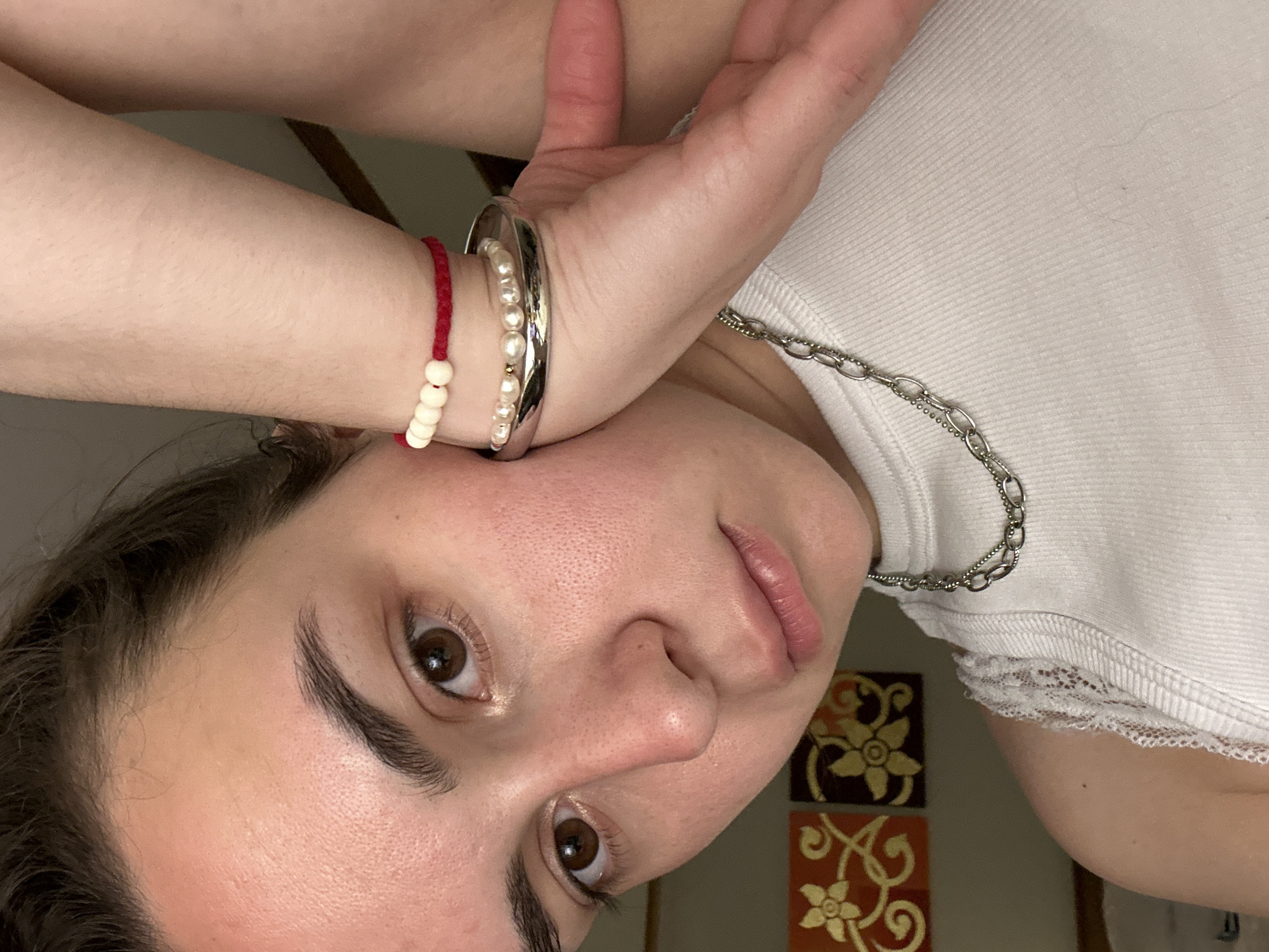 A medium, close-up shot depicts a person with fair skin and brown eyes. They wear a white tank top with lace trim. A silver chain necklace is visible. The person's left arm, adorned with three bracelets (a red string with white beads, a pearl bracelet, and a silver bangle), is positioned near their face. In the background, two decorative wall tiles featuring floral designs can be seen. The setting is indoor with warm lighting. There is no indication in the image to determine that the location is in Pattaya City, Thailand.