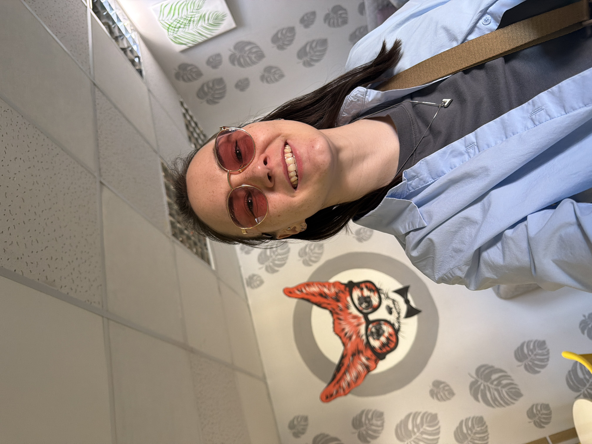 A smiling woman wearing red-tinted glasses, a gray shirt, and a light blue overshirt poses in an indoor setting. She is also wearing a brown strap across her chest. The background is a wall decorated with stylized leaf patterns and a circular graphic featuring a character with oversized glasses and red, wing-like shapes. The location appears to be in a building in the city of Chaikovsky, Russia. The ceiling has a textured, tiled appearance.
