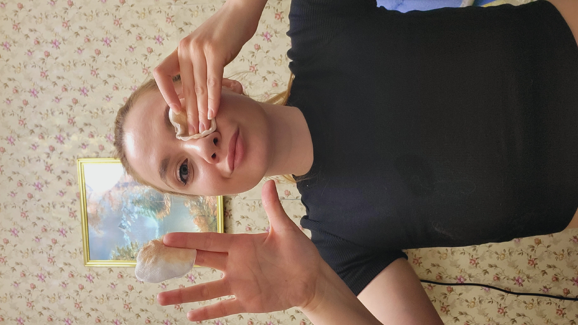 A young woman is shown from a low-angle perspective. She is wearing a black shirt and is holding two cotton pads, one near her eye and the other in her outstretched hand. The cotton pad near her eye appears to be clean, while the one in her hand is soiled with what appears to be makeup residue. The background consists of a floral patterned wallpaper and a framed picture of a landscape. The scene is indoors. There is no evidence to indicate that the image was taken in the city of Зипуново, Russia.