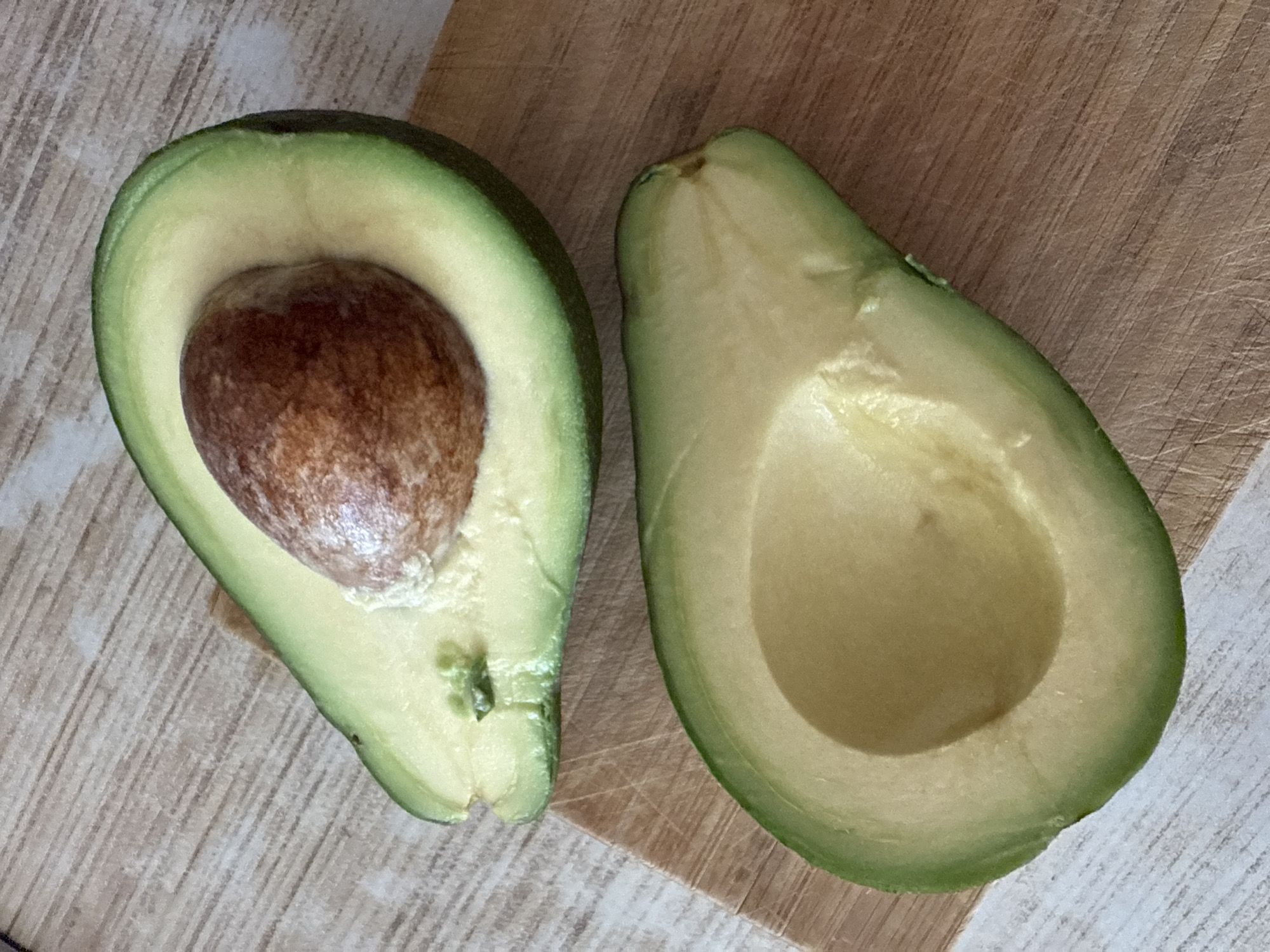 The media file displays a top-down view of two halves of an avocado on a wooden surface. The avocado on the left is whole and still contains its large brown pit, while the avocado on the right has been halved, revealing the smooth, pale green flesh and the empty cavity where the pit would have been. The avocados have a dark green skin with some lighter green undertones. The background is a light-colored wooden surface with a subtle wood grain pattern. To the right of the avocados, a distinct wooden cutting board with a more pronounced grain and a slightly darker hue is visible, suggesting the avocados have been prepared or are about to be prepared. The lighting appears to be natural, casting soft shadows. No people are present in the image, and the focus is solely on the avocados and their immediate surroundings. The scene is simple and domestic, likely a kitchen setting. There is no visible text in the image. The location context provided is Kavkazkaya, Russia.