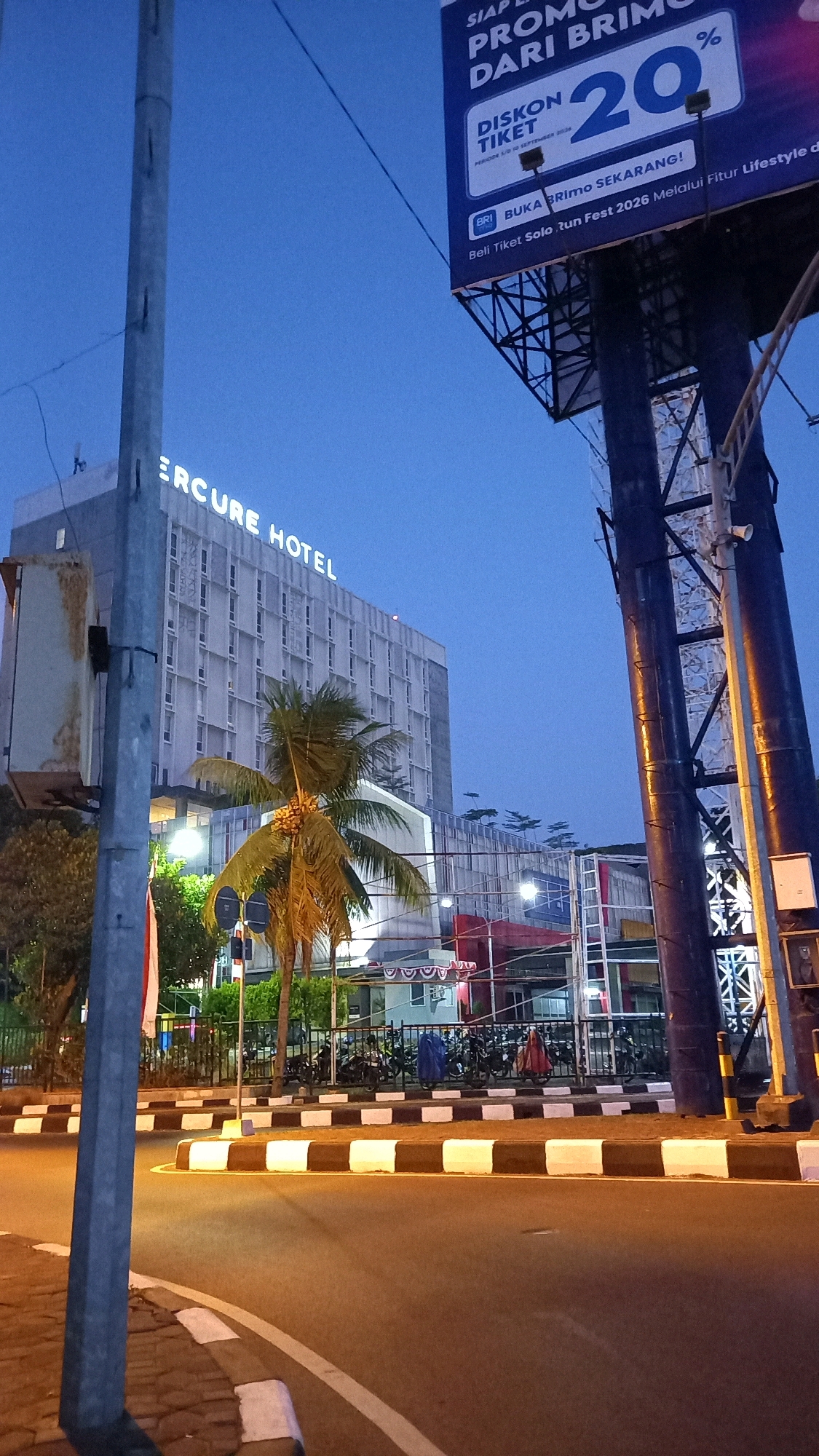 The image depicts a street scene in Surakarta, Indonesia, at dusk. A tall, modern building with "MERCURE HOTEL" illuminated in white lettering is prominent on the left side. In front of the hotel, a palm tree with a cluster of yellow coconuts stands. To the right of the hotel, a large billboard displays an advertisement for a "20% DISKON TIKET" for the "BRImo" application, with text indicating a promotion period ending September 2026. The billboard is supported by dark blue, industrial-looking structures. The street in the foreground features a paved road with white and brown striped curbs, indicating a traffic island or pedestrian crossing. Several motorcycles are parked behind a black metal fence in the middle ground, suggesting a public area or parking lot. The sky is a deep blue, characteristic of twilight. A concrete pole with electrical equipment is on the far left, and a traffic light is visible behind the palm tree. The overall atmosphere is urban and commercial.