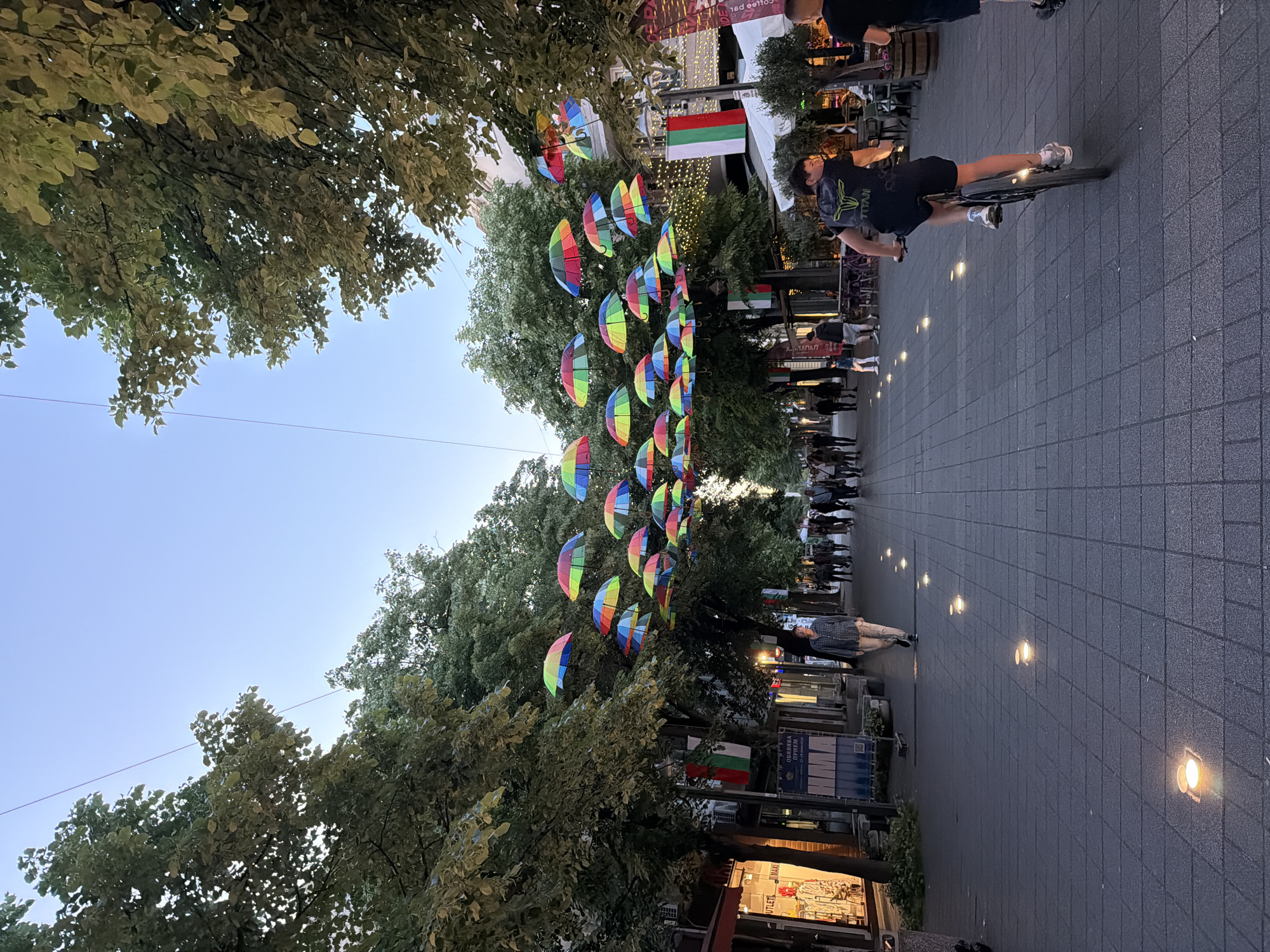 Captured at an oblique angle, the scene unfolds on a pedestrian street in Burgas, Bulgaria. Above, a series of rainbow-colored umbrellas are suspended between trees, framing the street. Bulgarian flags are prominently displayed. A person rides a bicycle through the center of the walkway, alongside pedestrians moving in both directions. The street is paved with grey bricks and punctuated by embedded lights. Shops and buildings line the thoroughfare. The sky is pale blue.