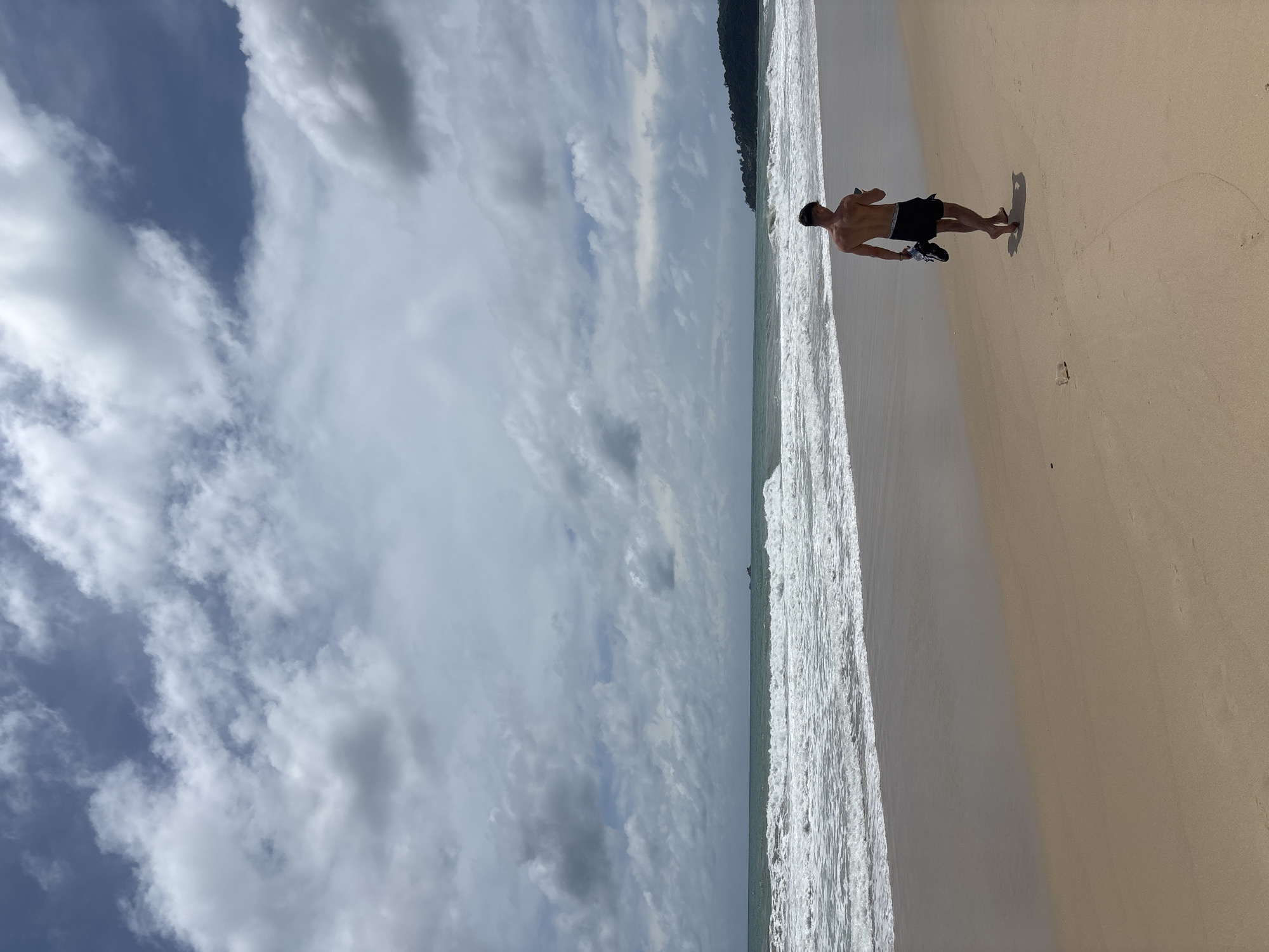 A shirtless man in black shorts walks barefoot along the sandy beach of Bang Tao in Choeng Thale, Thailand, carrying his shoes. The beach is broad and stretches far into the distance. The ocean waves roll onto the shore on the left side of the frame. The sky above is filled with white and gray clouds, and there's a glimpse of trees in the distance along the coastline.
