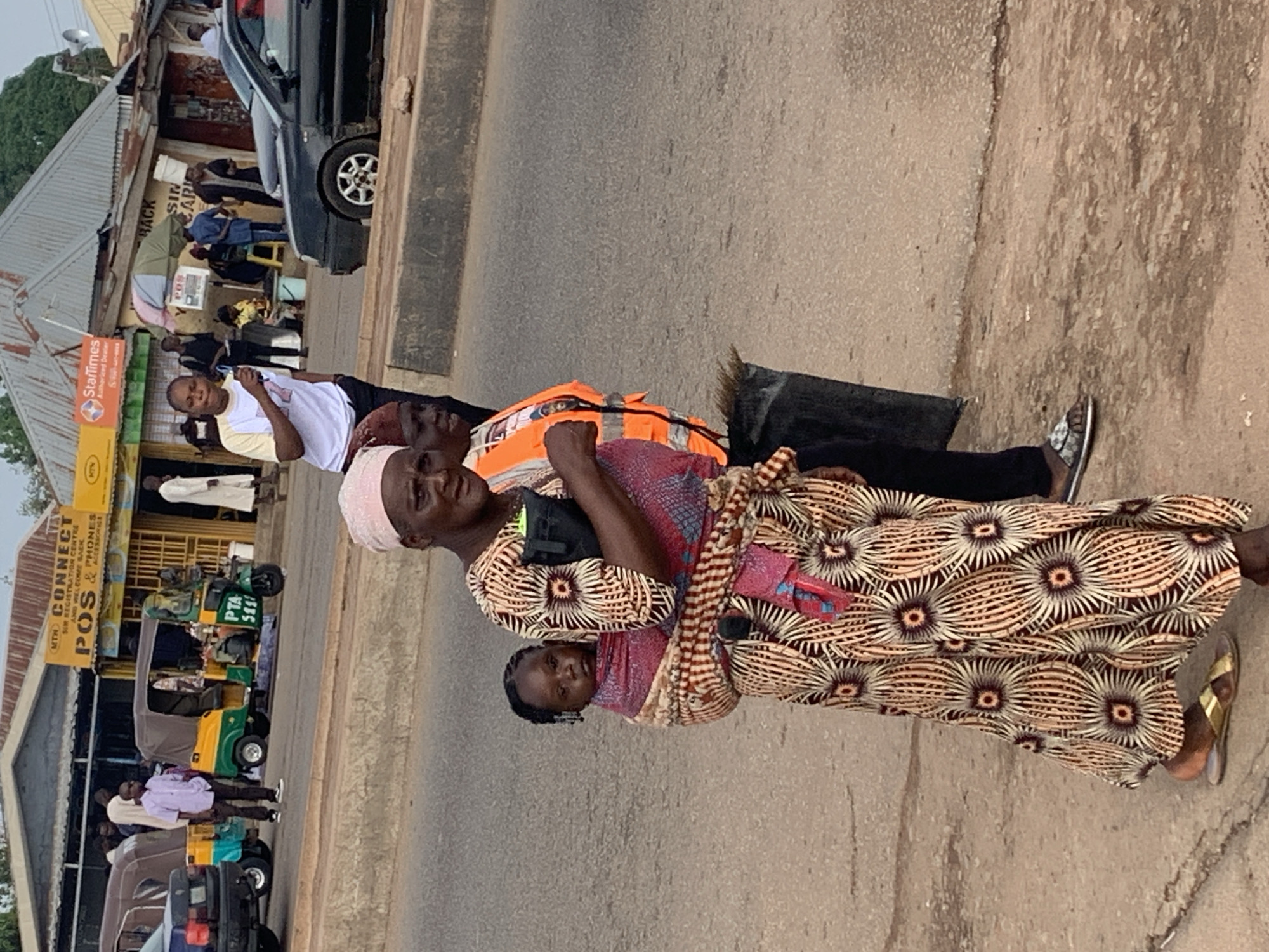 The image depicts a street scene in Gura Top, Nigeria. In the foreground, a woman wearing a patterned dress and headscarf carries a baby on her back, also wrapped in fabric. She is looking towards the left of the frame. Beside her, another person, possibly a woman, is wearing an orange safety vest. A large black bag rests on the ground near them. In the mid-ground, several people are visible near shops. One man in a light purple shirt stands near a yellow and green auto-rickshaw. Behind him, another auto-rickshaw is parked. A black car is visible in the right mid-ground. The environment appears to be a roadside with a paved road and a dirt sidewalk. Signs for "MTN CONNECT POS & PHONES ACCESSORIES" and "StarTimes Authorized Dealer" are visible on the buildings. The sky is overcast, suggesting daytime.