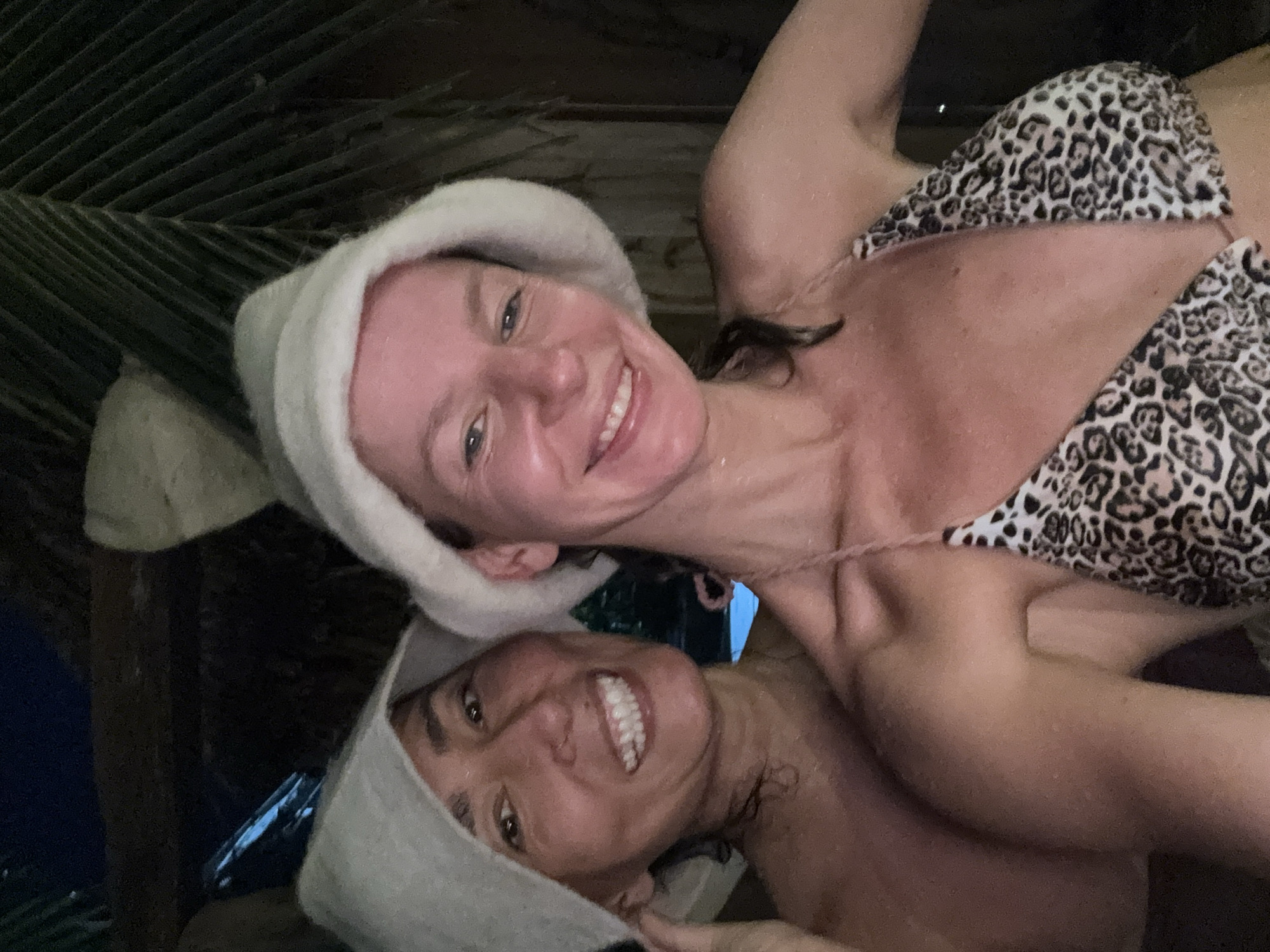 Two women, both wearing light-colored fuzzy hats, take a selfie in what appears to be a sauna or steam room. The woman in the foreground on the right is wearing a leopard-print bikini top, and her skin is glistening with moisture. The woman behind her on the left, also appearing to be wet, has her arm around the front woman and is smiling broadly. The background is dark, with some glimpses of what could be palm fronds and wooden structures, suggesting an outdoor or semi-outdoor location in a tropical environment. The lighting is dim, with a blue light source visible in the upper left quadrant. The setting is consistent with a spa or resort in Mũi Né, Vietnam, known for its beaches and wellness facilities.
