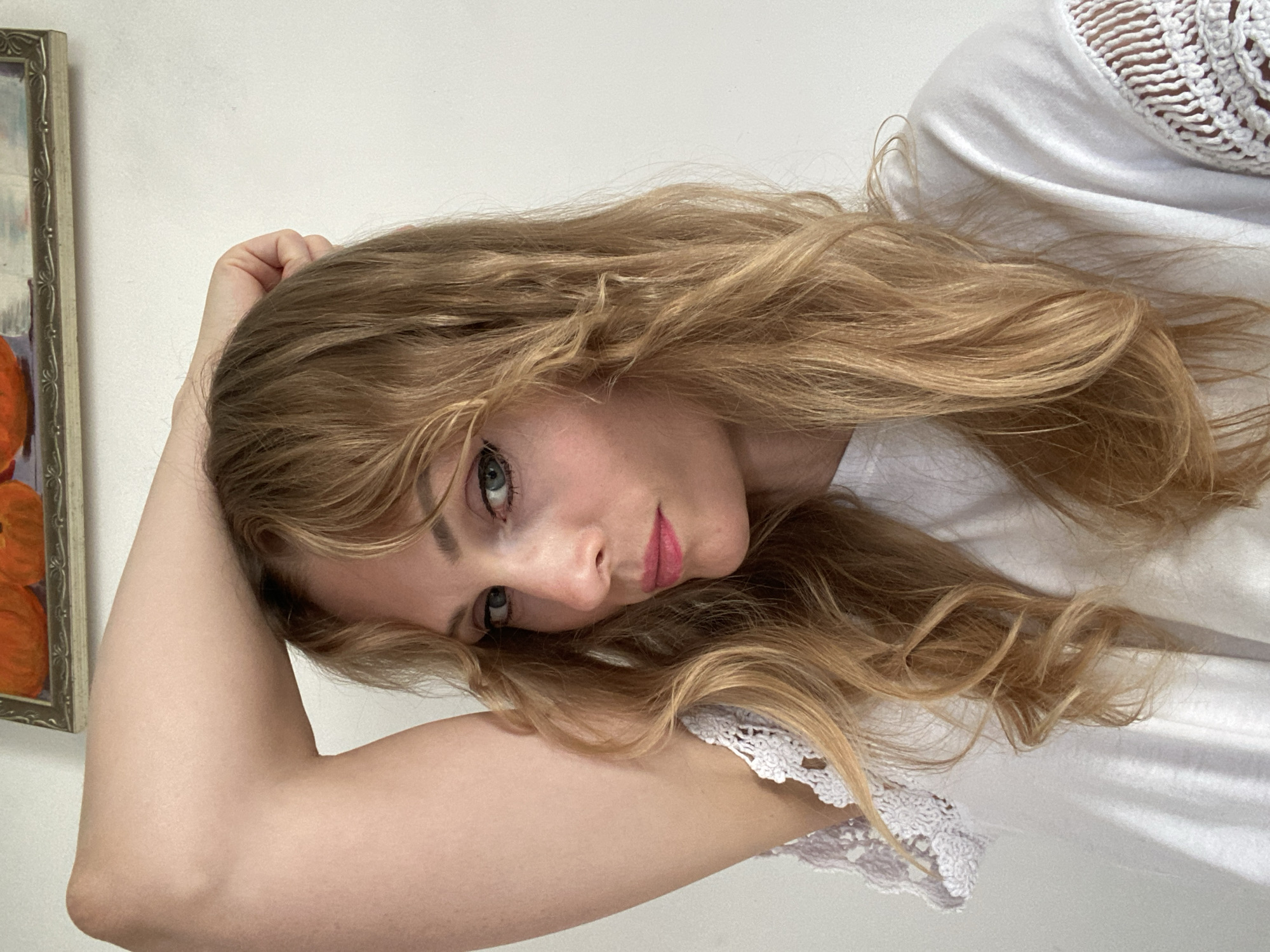 A woman is posing for a selfie with her arm raised behind her head. She has blonde, wavy hair and is wearing a white shirt with lace detailing on the sleeves. She is wearing pink lipstick and has eyeliner on. In the background, a framed painting of pumpkins is visible on a white wall. The image appears to be taken indoors, in a room with plain white walls. There is no visible indication of the location being in Міханавіцкі сельскі Савет, Belarus.