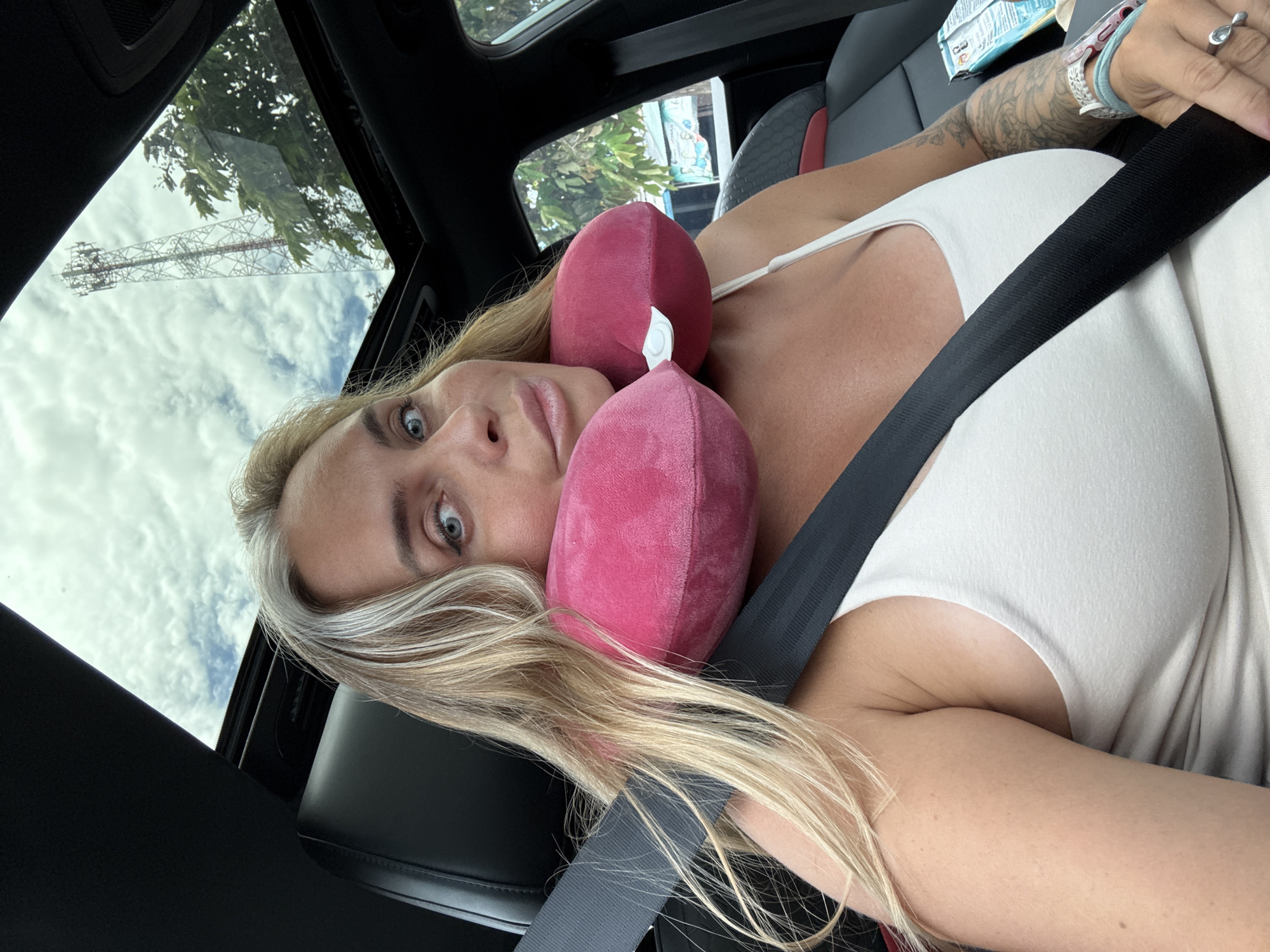 A woman with blonde hair, streaked with grey, is seated in a car. She is wearing a white tank top and a pink neck pillow. A black seatbelt is fastened across her chest. Her left arm is resting on the seatbelt, and her hand is visible with rings on her fingers and a teal bracelet. A package of something is visible on the car seat to her right. The car has a sunroof open, revealing a cloudy sky with a tall communication tower and green trees visible. The scene appears to be in Tambon Pak Phraek, Thailand, suggested by the presence of the communication tower and lush vegetation typical of the region.