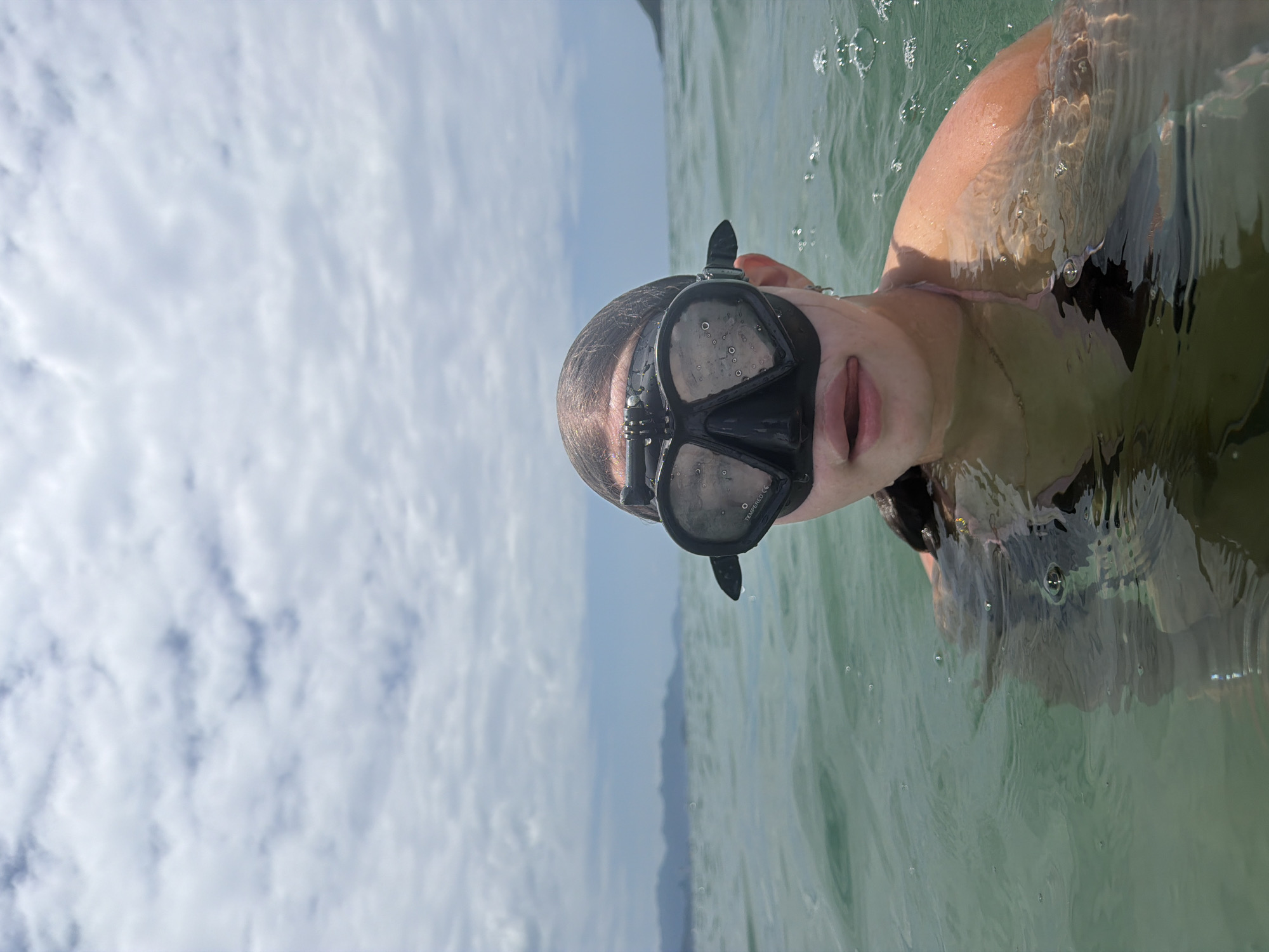 A person wearing a black diving mask and a pink bikini strap is in the ocean, with their head and shoulders visible above the water. The water is a greenish hue, and there are ripples and some small bubbles on the surface. The sky above is cloudy with a few patches of blue. In the distance, mountains and a coastline are visible. The scene appears to be a sunny day. The location is likely a beach or bay in Nha Trang, Vietnam, given the presence of ocean, mountains, and a clear sky indicative of a tropical climate.
