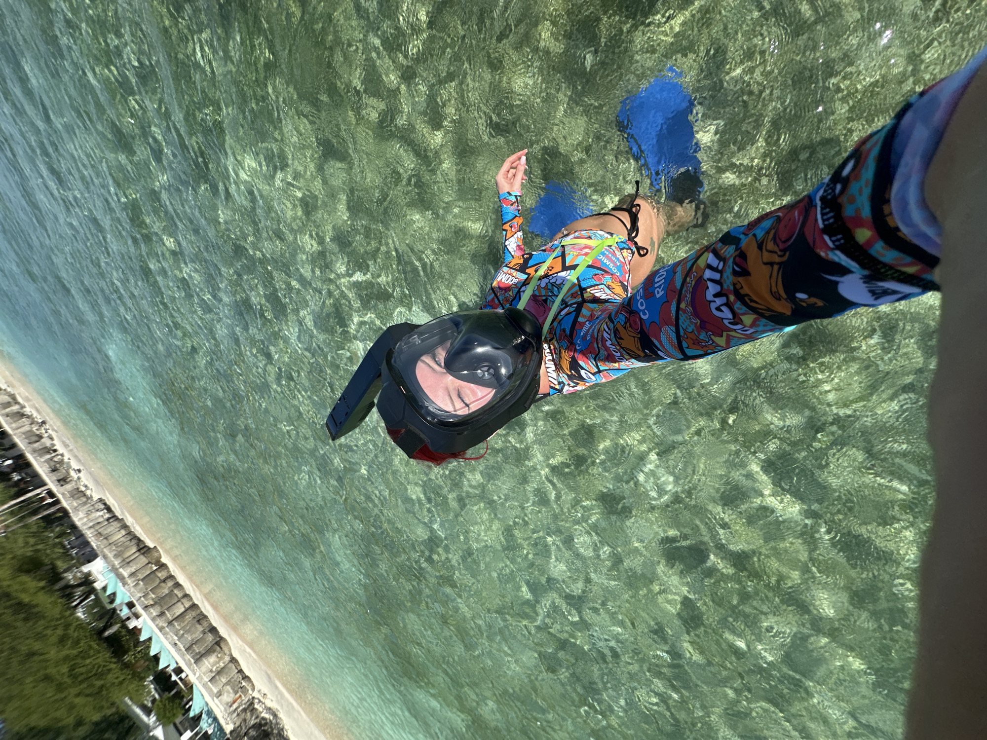 A woman floats in clear, shallow water, wearing a full-face snorkel mask, colorful, long-sleeved rash guard, bikini bottoms, and blue fins. The scene appears to be near a beach in Unknown, Indonesia. Behind the woman, there's a sandy beach with beach chairs, umbrellas, and foliage. The water is clear, showing the sandy bottom. The woman is holding the camera out to take a selfie.