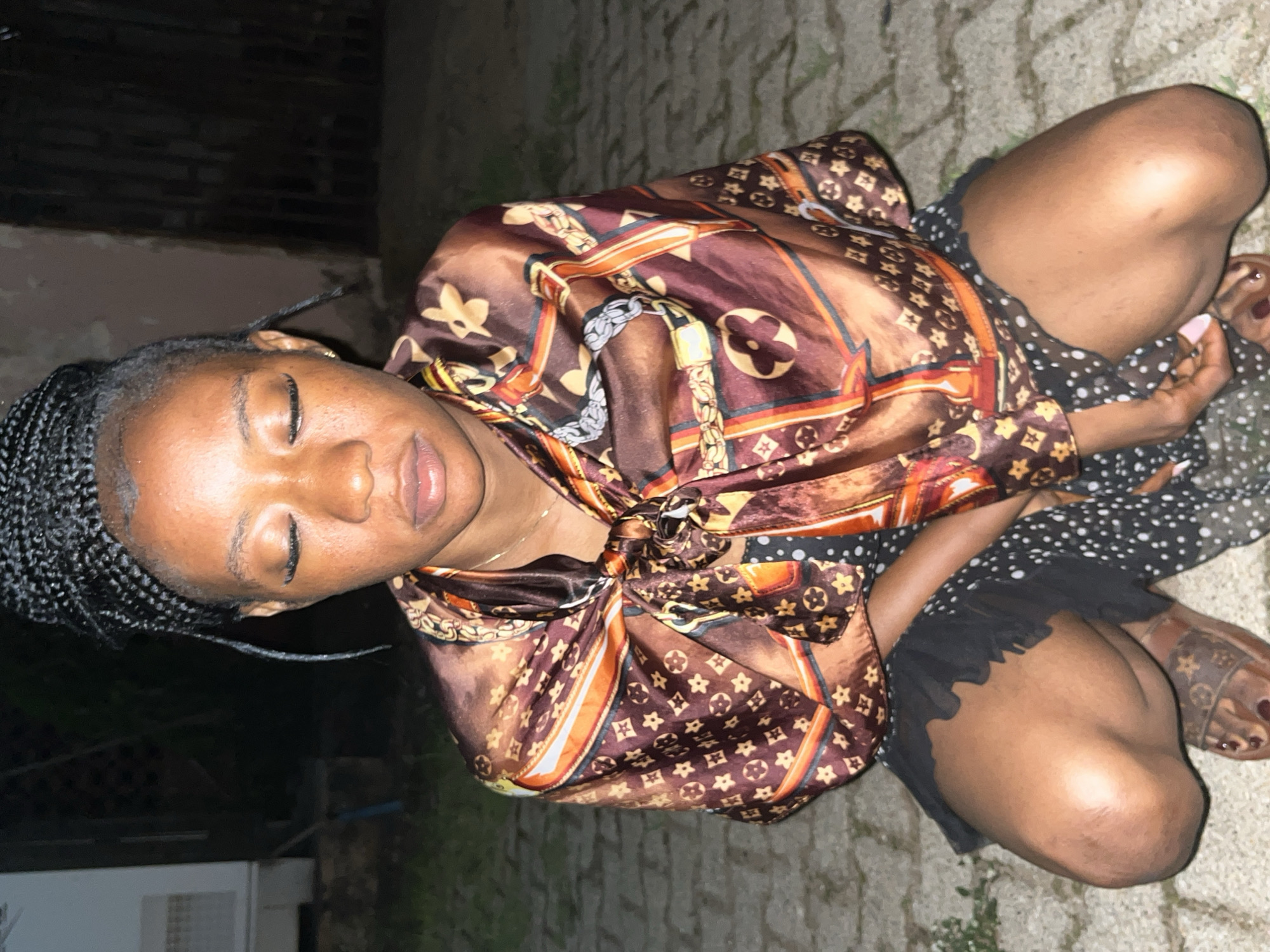 The image shows a young Black woman squatting on a paved surface, possibly an outdoor area in the city of Jos, Nigeria. She has her eyes closed and her head bowed slightly. Her hair is styled in braids that are pulled up into a bun. She is wearing a patterned silk scarf tied around her neck, a black polka-dot dress, and brown sandals with a repeating monogram pattern. The background is dark and out of focus, with hints of buildings and foliage. The lighting suggests it is nighttime or dusk.