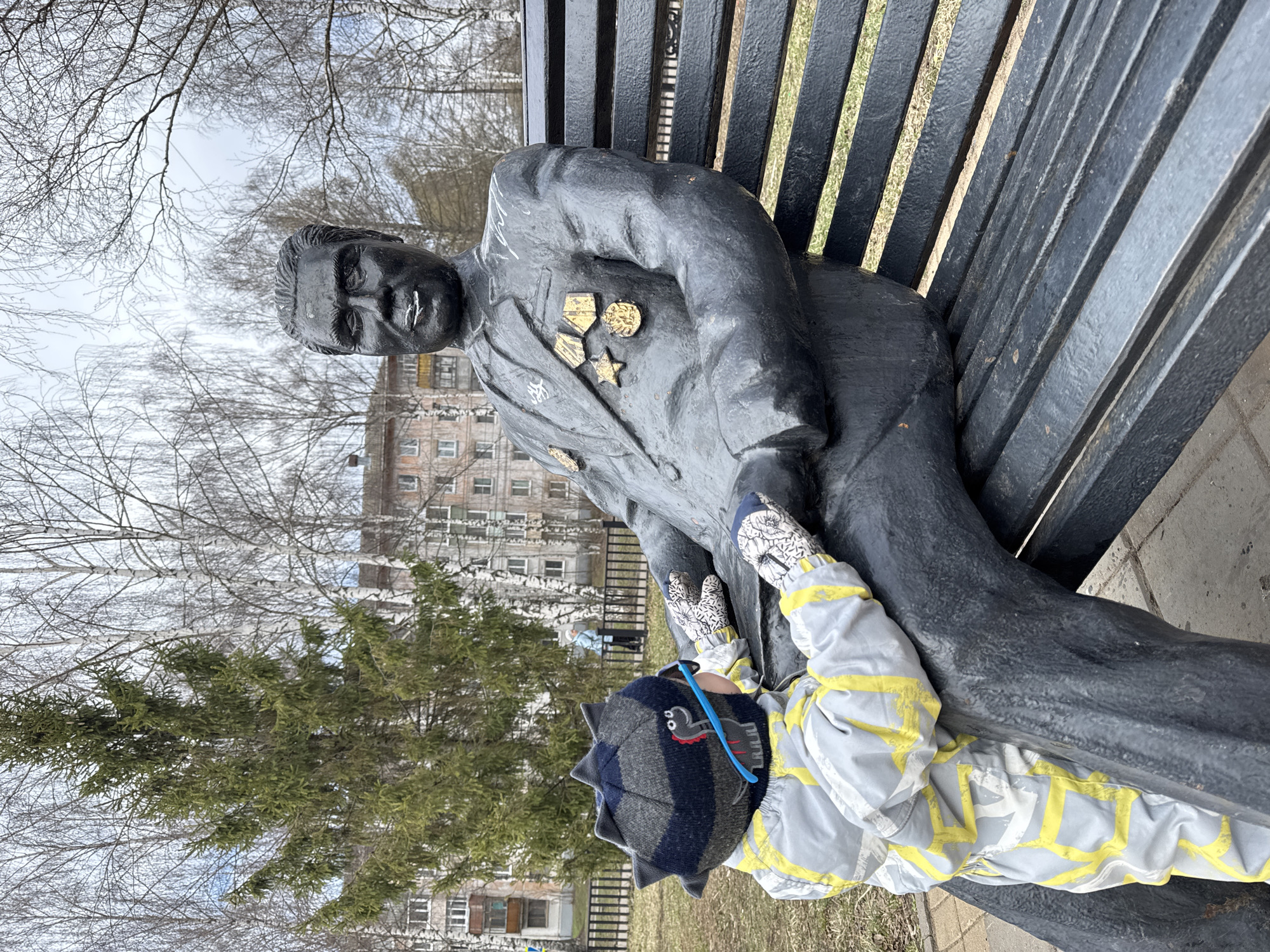 A statue of a man in a suit sits on a black metal park bench. Medals are pinned to the left side of the statue's suit. A child in a gray and yellow jumpsuit lies on the ground, with their hands reaching up and touching the statue. The child is wearing a hat with cartoon dinosaur embellishments. Behind the statue is a green pine tree, bare trees, a multistory building, and a cloudy sky. The statue is of Kuzebay Gerd, a Udmurt poet, ethnographer, and playwright, located on Sovetskaya Street in Izhevsk, Russia.