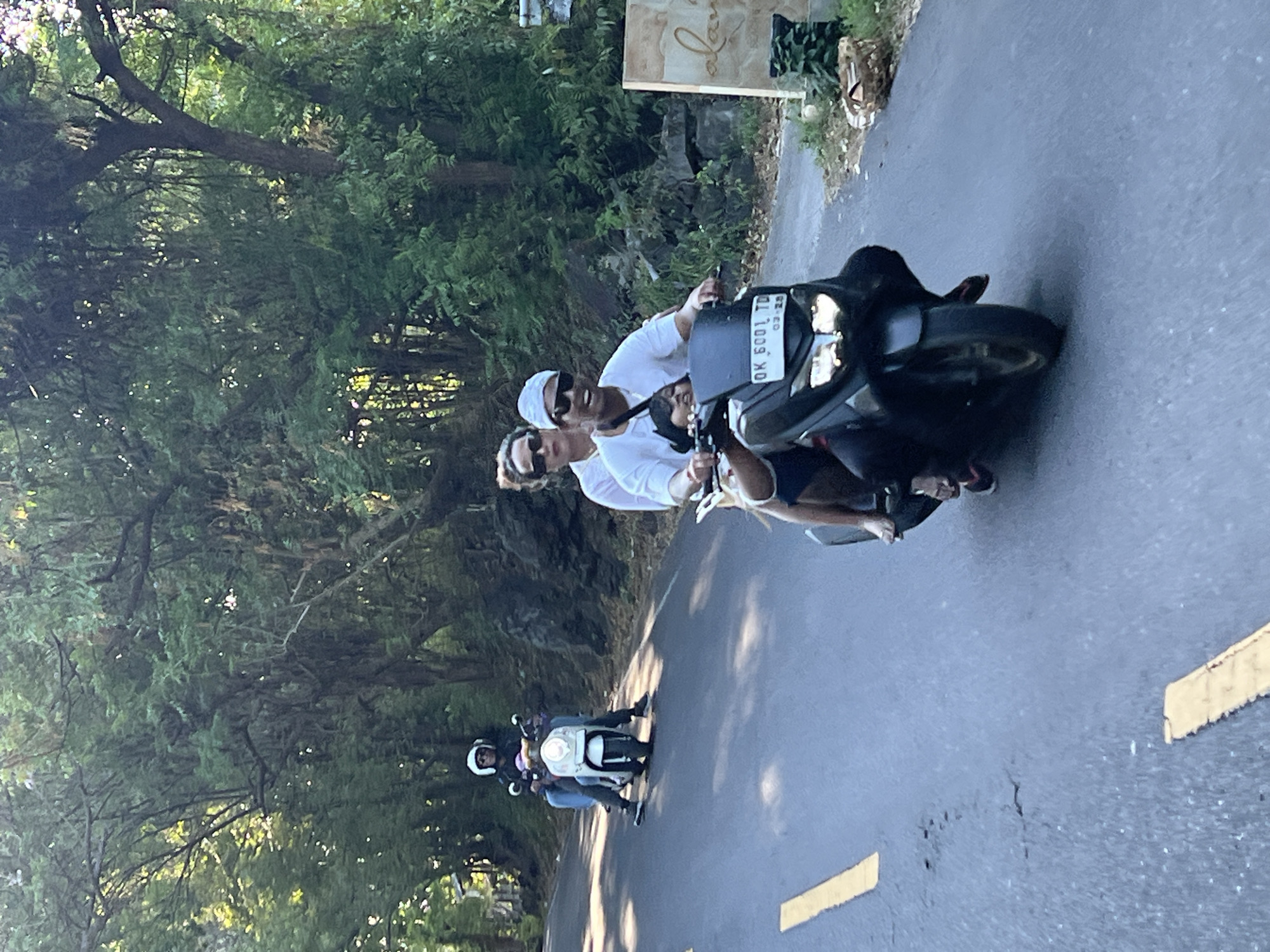 In Kubu, Indonesia, on a paved road surrounded by lush green foliage and trees, three people are riding a scooter. A man, wearing a white t-shirt, sunglasses, and a white head covering, is driving the scooter. A woman, with sunglasses and wearing a white top, is riding behind him. A child is seated in front of the man. Another scooter, a white one, is visible in the background with two individuals riding it. The road has yellow markings. A sign with the word "alani" is visible on the right side of the frame.