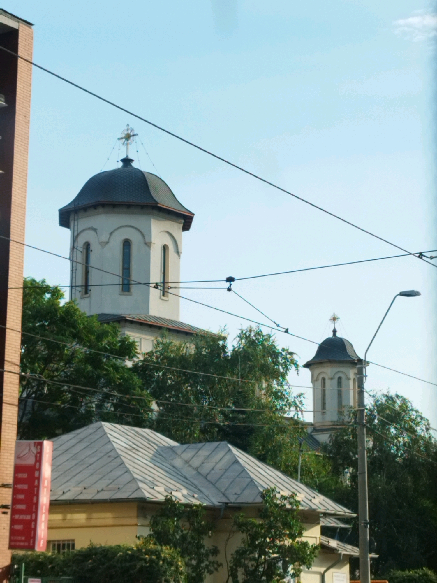 The image depicts a view of buildings in Bucharest, Romania, under a clear blue sky. In the foreground, a building with a light yellow facade and a gray, gabled metal roof is visible. To the left, a tall, narrow brick building stands out. In the background, two church towers with dark, domed roofs are prominent. The larger tower on the left is octagonal and white, with arched windows and a cross on top. The smaller tower on the right is also white with arched windows and a cross. Overhead, power lines crisscross the sky, obscuring parts of the scene. Dense green trees are scattered throughout the image, particularly around the bases of the churches and behind the foreground building. A red sign with white text, appearing to advertise dental services, is partially visible on the left. The overall impression is an urban landscape with religious architecture amidst trees and infrastructure.
