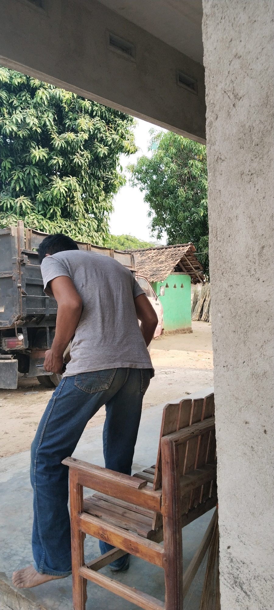 The image depicts an outdoor scene in Tanjung Ratu Ilir, Indonesia, under bright daylight. In the foreground, a person wearing a grey t-shirt and blue jeans is partially visible, bending over, with their back to the camera. They are barefoot on a concrete floor. To the right of the person, a weathered wooden chair is positioned, facing partially away from the viewer. Behind the person, a dark-colored truck bed is visible. In the background, a small, bright green building with a tiled roof is situated. Lush green trees with dense foliage dominate the upper portion of the image, suggesting a rural or semi-rural environment. The overall impression is one of a casual, everyday moment in a tropical setting.
