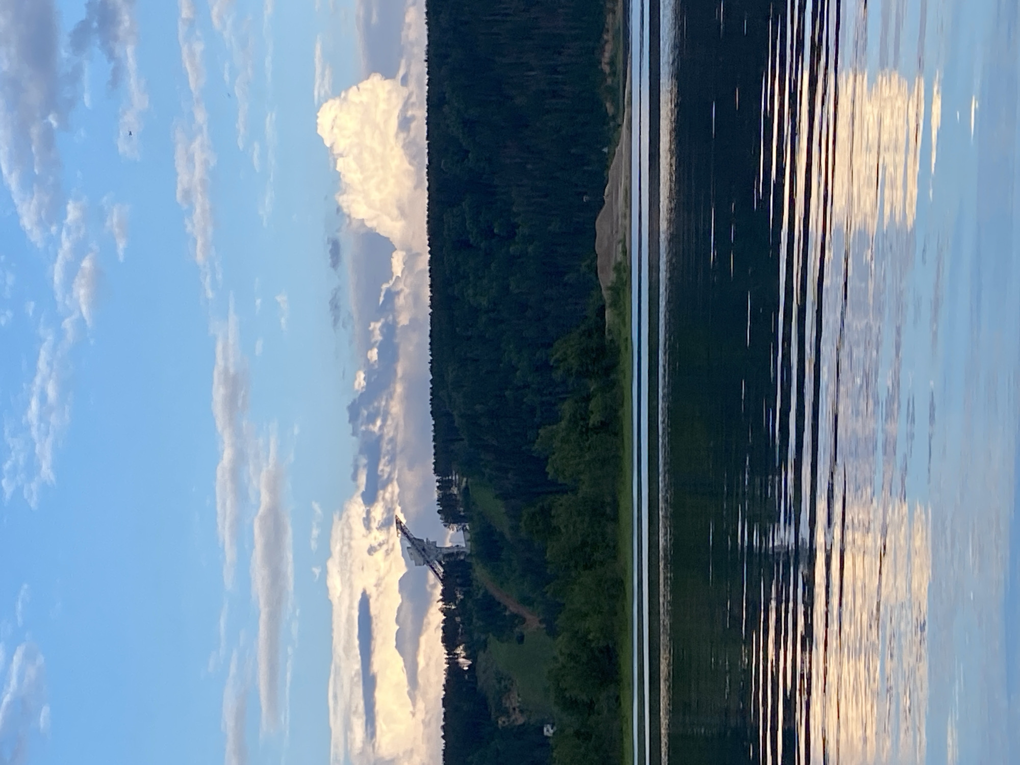 The image depicts a serene landscape with a large body of water in the foreground, reflecting the sky and surrounding scenery. A dark, dense forest covers a hillside on the left side of the frame, transitioning into a lighter, more rugged terrain higher up. A prominent man-made structure, resembling a tower or a large antenna, is visible on the hillside amidst the greenery. The sky is a mix of blue and white, with scattered clouds, some of which are illuminated by sunlight, suggesting it might be late afternoon or early evening. The water's surface is disturbed by ripples, creating elongated reflections of the sky and the structure on the shore. The overall mood is tranquil and natural, with the man-made element contrasting with the wild landscape. The location context provided is Noviy, Russia.