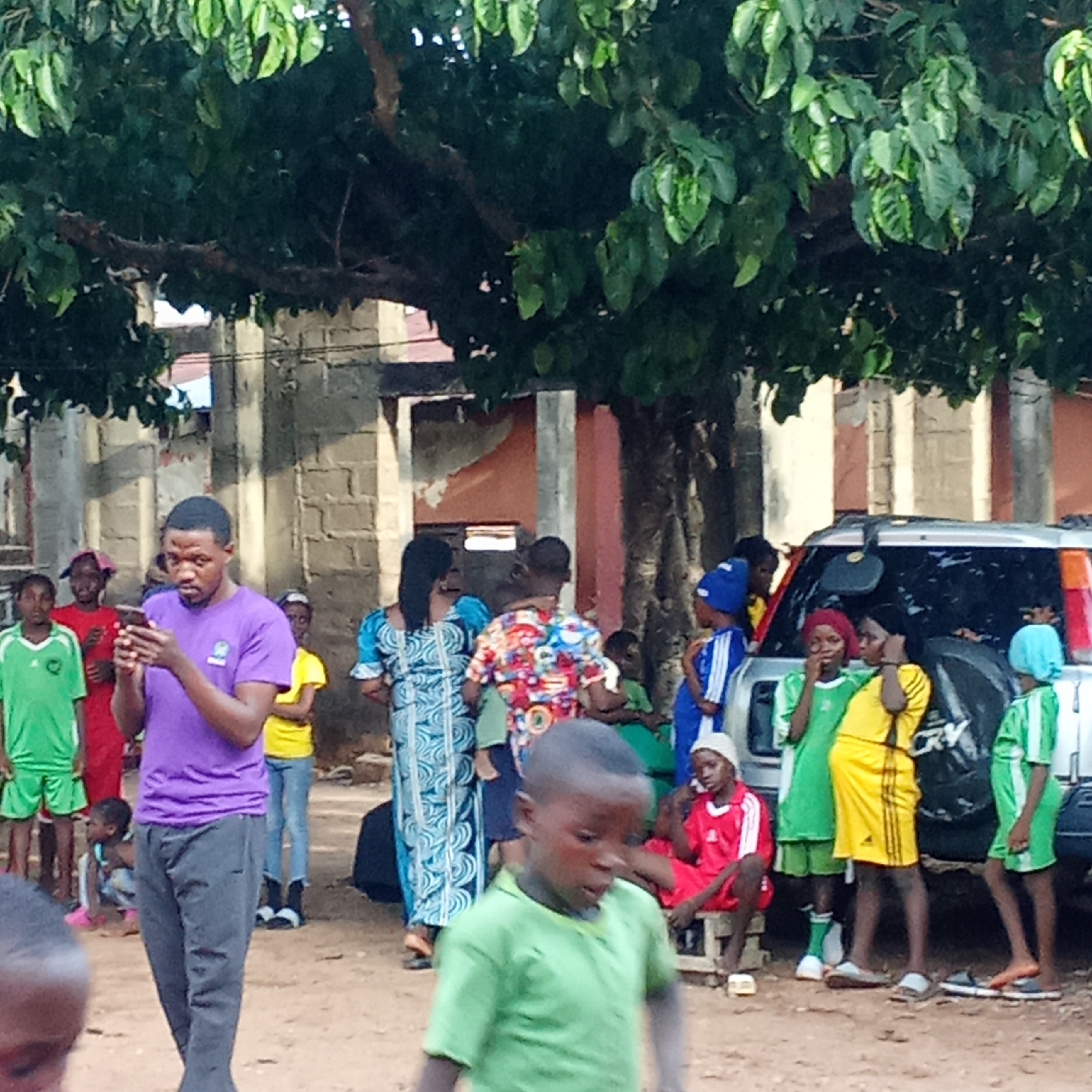 The image shows a group of people in Akwanga, Nigeria, standing outside near a building and under a large leafy tree. A man in a purple shirt is looking at a mobile phone in his hand. Several children are wearing sports uniforms in colors of green, yellow, and red. A silver SUV is parked nearby. A woman in a blue patterned dress is standing with a man in a colorful shirt holding a child. Other people are present in the background.