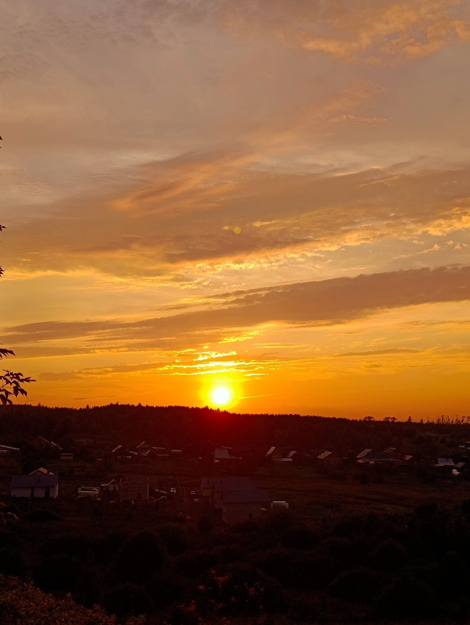 The image captures a sunset over a rural settlement that appears to be located in the vicinity of Фоки, Russia. The sky is dominated by vibrant orange and yellow hues, with elongated, wispy clouds stretching across the horizon. The sun, a bright disc of light, is low in the sky, casting a warm glow over the landscape. In the foreground, silhouetted against the vibrant sky, is a dark, undulating treeline. Below the treeline, nestled within the landscape, are several low-rise buildings, characteristic of a village or small settlement. These structures are mostly dark and indistinct due to the low light conditions, but some appear to have roofs with varying pitches. The ground between the buildings is covered in vegetation, also appearing dark and dense. The overall impression is one of a tranquil, rural evening scene. There are no people or discernible actions visible in the image.