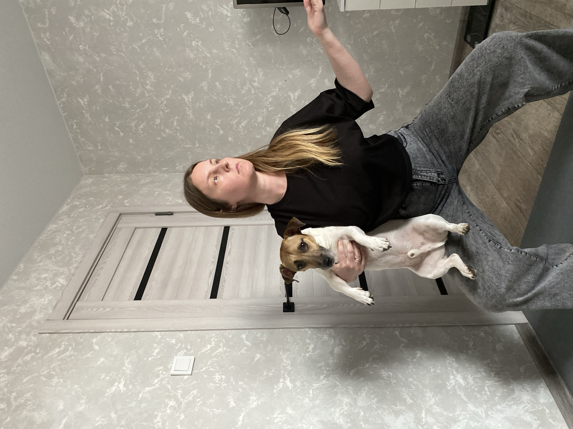 A woman stands in a room holding a Jack Russell Terrier. The woman has blonde hair and wears a black shirt and gray jeans. The dog is primarily white with brown markings on its head and ears. The room has gray, textured walls, a light-colored door with dark vertical lines, and what appears to be a switch on the wall near the door. There is a dark-colored electronic device mounted high on the wall. The location is likely indoors in a home or apartment, but I am unable to determine if the location is in the city of Noviy, Russia.