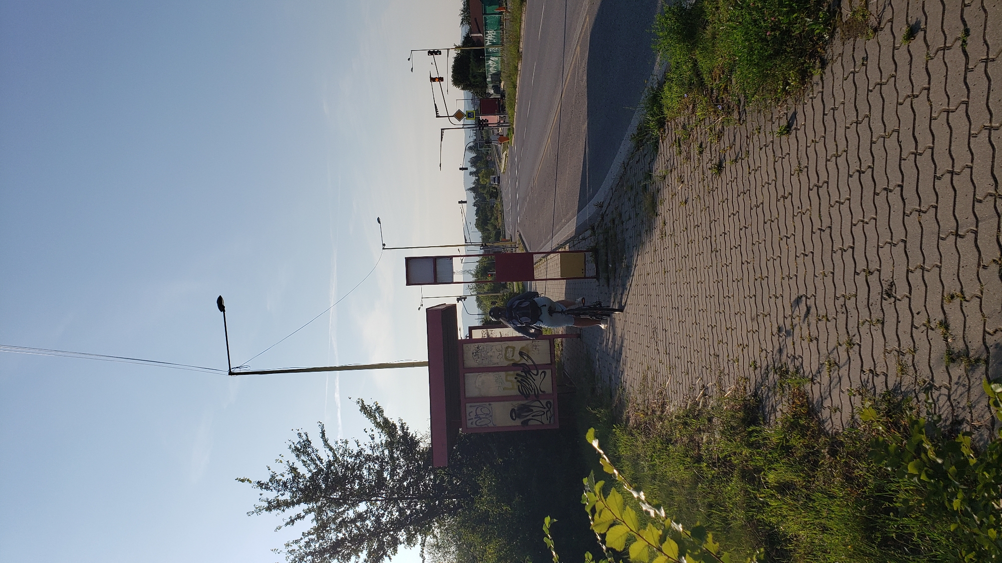 The image shows a person riding a bicycle on a brick pathway next to a bus stop. The bus stop is a red and silver structure with graffiti on its panels. The pathway is lined with green grass and some weeds. To the right of the pathway is a paved road with traffic lights and a few vehicles. In the background, there are trees and hills, suggesting a suburban or semi-rural setting. The sky is clear and blue, indicating a daytime scene. The bus stop is located on a street that appears to be a main road, possibly with a junction or intersection further down. The overall scene depicts a typical urban or suburban environment during daylight.
While it is impossible to pinpoint the exact location in Prešov, Slovakia, from the image alone, the presence of a bus stop, paved roads, traffic lights, and a somewhat developed landscape suggests it is an accessible area within the city or its immediate surroundings. The style of the bus stop structure and the road infrastructure do not immediately identify a specific, well-known landmark within Prešov.