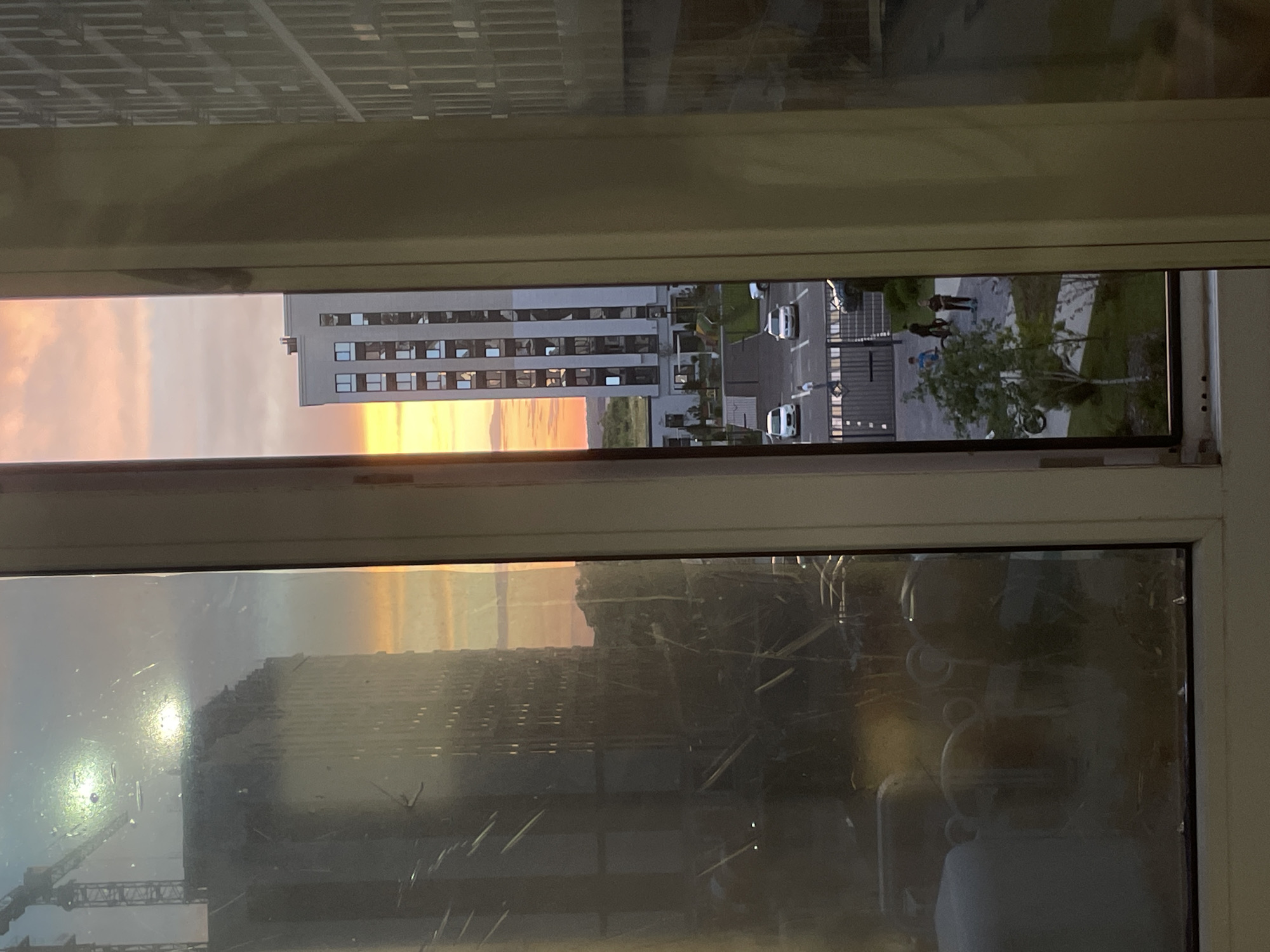 This is a photograph taken from a window in a multi-story building, likely a residential apartment, in Stavropol, Russia. The view looks out onto an urban landscape characterized by modern architecture and a sunset.
In the foreground, the window frame divides the image into two vertical sections. The left section is dominated by the interior of the room, showing reflections on the glass, including what appear to be household objects like containers or furniture, obscured by the window's condition, which shows streaks and smudges. A tall construction crane and the upper portion of a building under construction are visible through the window on the left, partially obscured by the reflections.
The right section of the window reveals the exterior scene. A modern, tall residential building with many windows is the central feature. A parking lot with several cars is visible in front of this building, along with a few people walking or standing. A small green area with trees is also present. Beyond the buildings, the sky displays a gradient of colors associated with sunset, with shades of yellow, orange, and pink. In the distance, more residential buildings and possibly some natural landscape are visible. The overall impression is a view of a developing urban area during the golden hour.