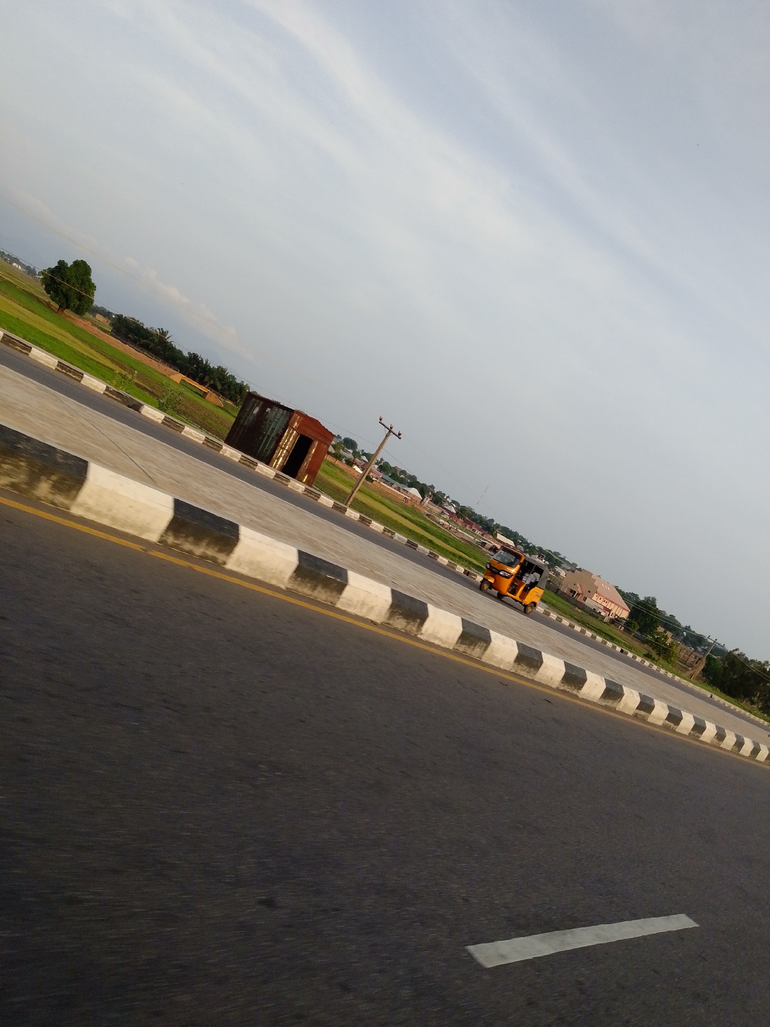 The image depicts a roadside scene in Jalingo, Nigeria. The foreground features a section of an asphalt road with a white lane marker and a concrete barrier with black and white diagonal markings. To the left of the road, there's a grassy field with a lone tree and what appears to be cultivated land with some structures in the distance. On the right side of the road, a dilapidated, rusty metal structure stands near the roadside. Further down the road, an orange auto-rickshaw, also known as a "Keke," is visible, carrying passengers. In the background, there are buildings, power poles, and more green landscape, suggesting a semi-urban or peri-urban environment. The sky is overcast with light clouds.