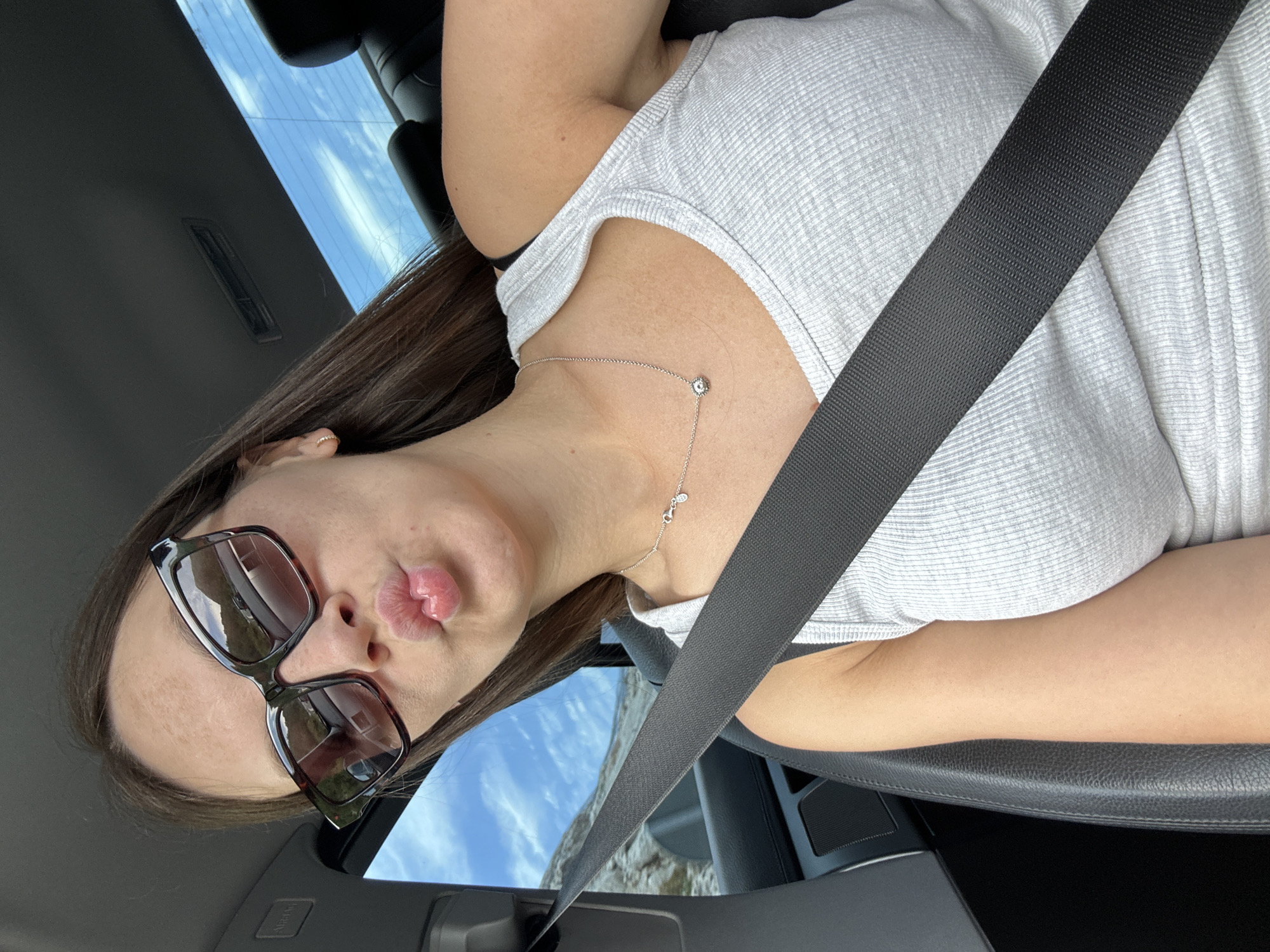A young woman with long brown hair and sunglasses is in the passenger seat of a car. She is wearing a grey tank top and a necklace. A seatbelt is across her chest. The background shows a blue sky with clouds and a rocky landscape. The image was taken in Ugnji, Montenegro.