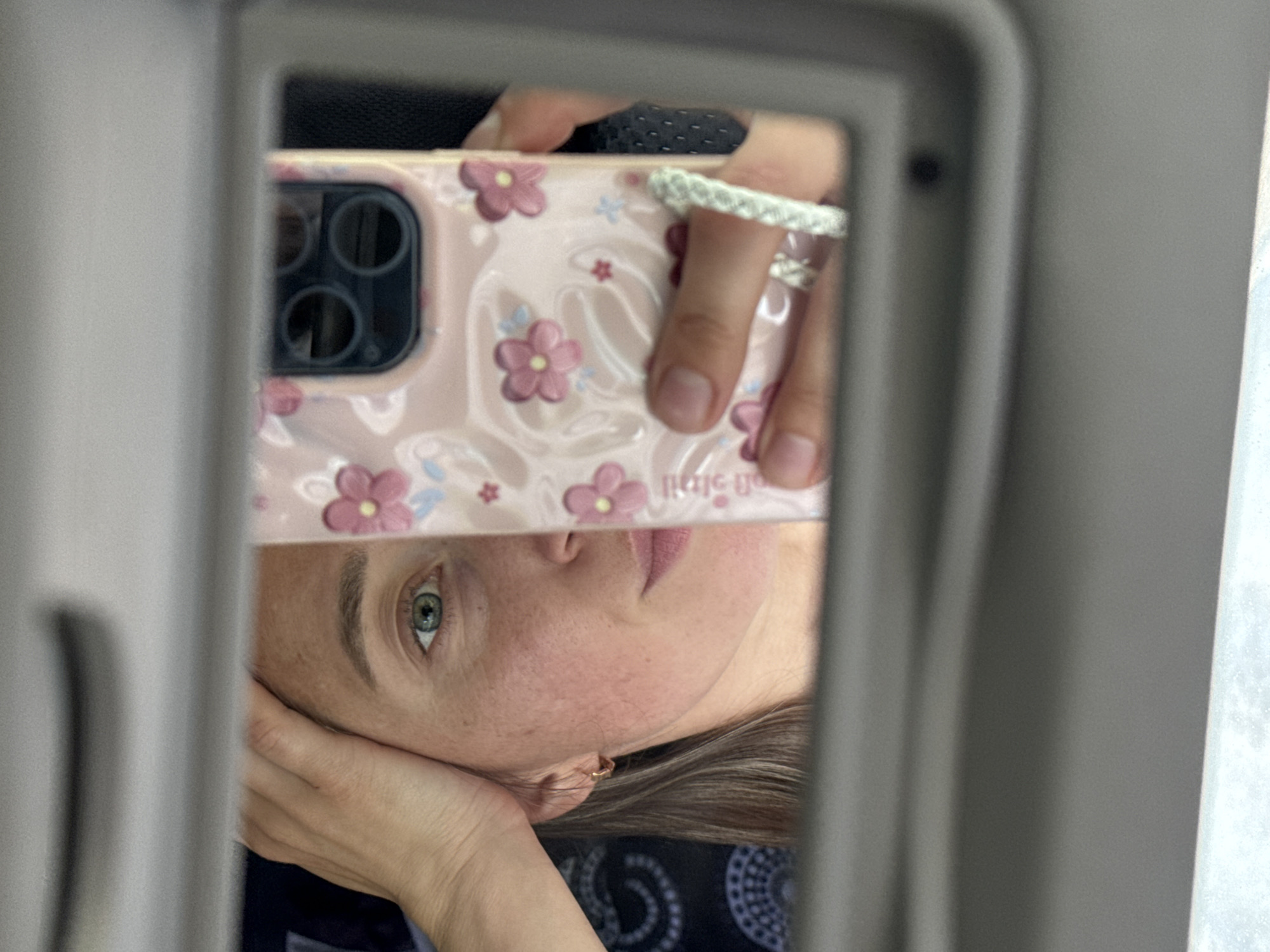 A self-portrait captured via reflection in a car's vanity mirror in Karagay, Russia. The image features the upper face of an individual, presenting a greenish-blue left eye, a defined eyebrow, and partial views of the nose, mouth, and a freckled cheek. The subject's right hand is positioned on the forehead. A small gold-colored hoop earring is visible in the left ear. A light pink smartphone, featuring a multi-lens camera array and encased in a floral-patterned "Casetify" case, is held vertically by the subject's left hand, obscuring a portion of the face. This hand displays a white braided ring on the middle finger and a second ring on the ring finger. The reflected background behind the subject's head consists of a dark fabric with a white circular pattern, consistent with vehicle interior upholstery. The grey frame of the sun visor mirror borders the reflective surface.