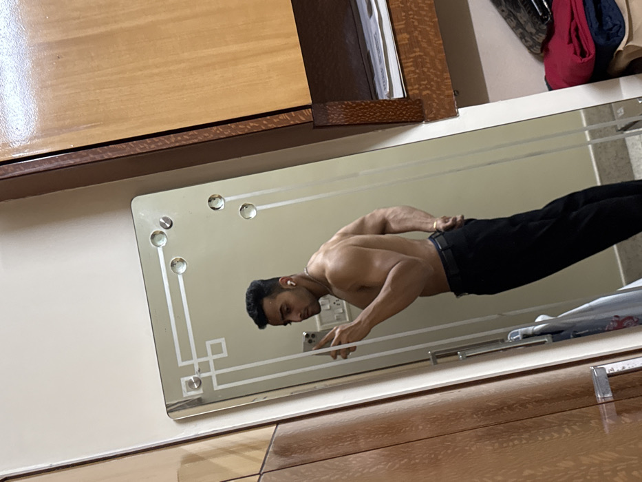 A shirtless man in black pants stands in front of a mirror, holding a phone. He has dark hair, a small beard, and wears a chain. The mirror is rectangular with decorative accents and is mounted on a wall. The man's reflection fills much of the mirror. The scene includes a wooden dresser top above the mirror and tiled flooring below. The lighting is soft, creating a slightly warm tone.