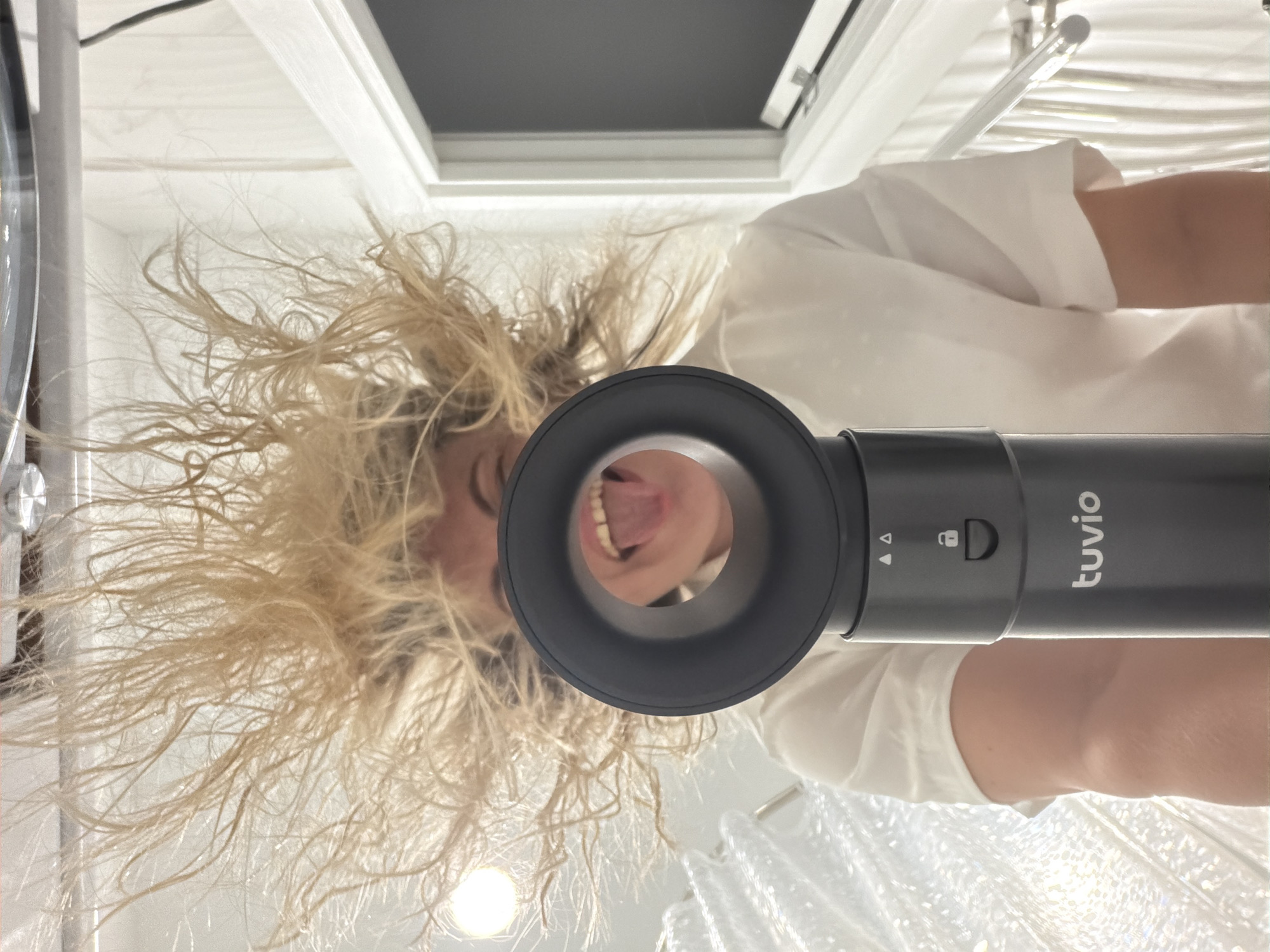 A low-angle perspective shows a person holding a "tuvio" hair dryer directly overhead. The person, visible through the dryer's opening, has disheveled, light blonde hair and is sticking out their tongue. They are wearing a white shirt. The background reveals a brightly lit room with a shower curtain and a small rectangular window. The exact location in Votkinsk, Russia cannot be determined from this image.