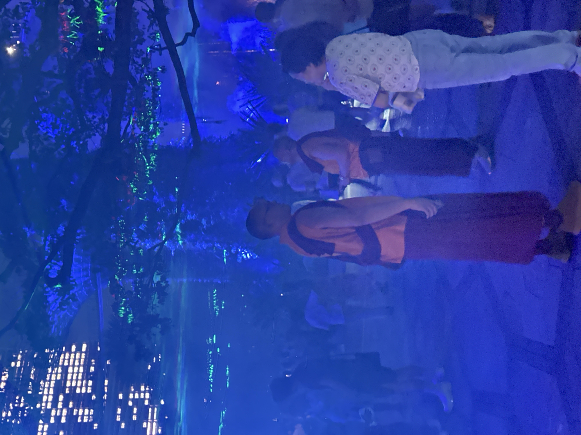 The image depicts a nighttime outdoor gathering or event, likely in Singapore, given the mention of the city. The environment is heavily illuminated with vibrant blue and green lights, creating a moody and atmospheric scene. In the foreground, three individuals are prominently visible. To the left, a person stands facing away from the camera, dressed in a reddish-brown robe and a dark vest or tunic. Their gaze is directed towards the right. In the center, another individual is seen from behind, also wearing a similar robe and a bright orange vest. This person appears to be looking to the right, possibly at the third individual. To the right, a person is depicted from the side, wearing a light-colored, patterned shirt and light blue pants. They are facing towards the left, their head slightly tilted down, and they are holding a white object, possibly a phone or a small bag.
In the background, numerous other people are scattered throughout the scene, appearing as blurred figures due to the low light conditions and the motion of the event. They are engaged in various activities, suggesting a lively and social atmosphere. The background also reveals architectural elements with bright, illuminated windows, possibly a building or a structure with many lights. Overhead, branches of trees are visible, also illuminated with blue and green lights, adding to the surreal and festive ambiance. The ground appears to be paved, with some visible patterns or textures. The overall impression is that of a dynamic event or festival, likely held in a public space in Singapore, with an emphasis on light installations and a celebratory mood.