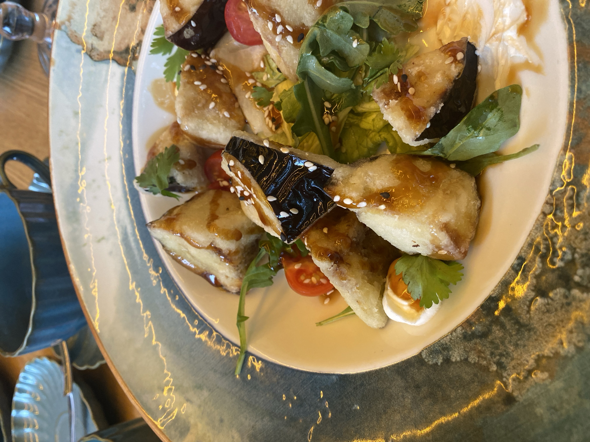The image shows a close-up, overhead view of a plate of food, likely a salad or appetizer. The main subjects are pieces of fried eggplant, which are golden brown and glazed with a dark sauce, sprinkled with sesame seeds. Interspersed with the eggplant are fresh green leaves of lettuce or arugula and halved cherry tomatoes. There is also a dollop of white sauce or dressing with a drizzle of sauce on top, garnished with a sprig of cilantro. The food is arranged on a decorative light-colored plate with an artistic, abstract pattern around the rim, featuring metallic gold accents. The plate is set on a dark, textured surface, possibly a table. In the background, hints of other objects are visible, including dark blue pottery or ceramic items and a wooden surface, suggesting a dining setting. The lighting appears to be warm and natural, possibly from a window, and the focus is entirely on the food. No people are visible, and there is no text. The overall impression is of a beautifully presented and appetizing dish. The location context provided is Tuymazy, Russia, which could imply this is a restaurant or home dining experience in that area.