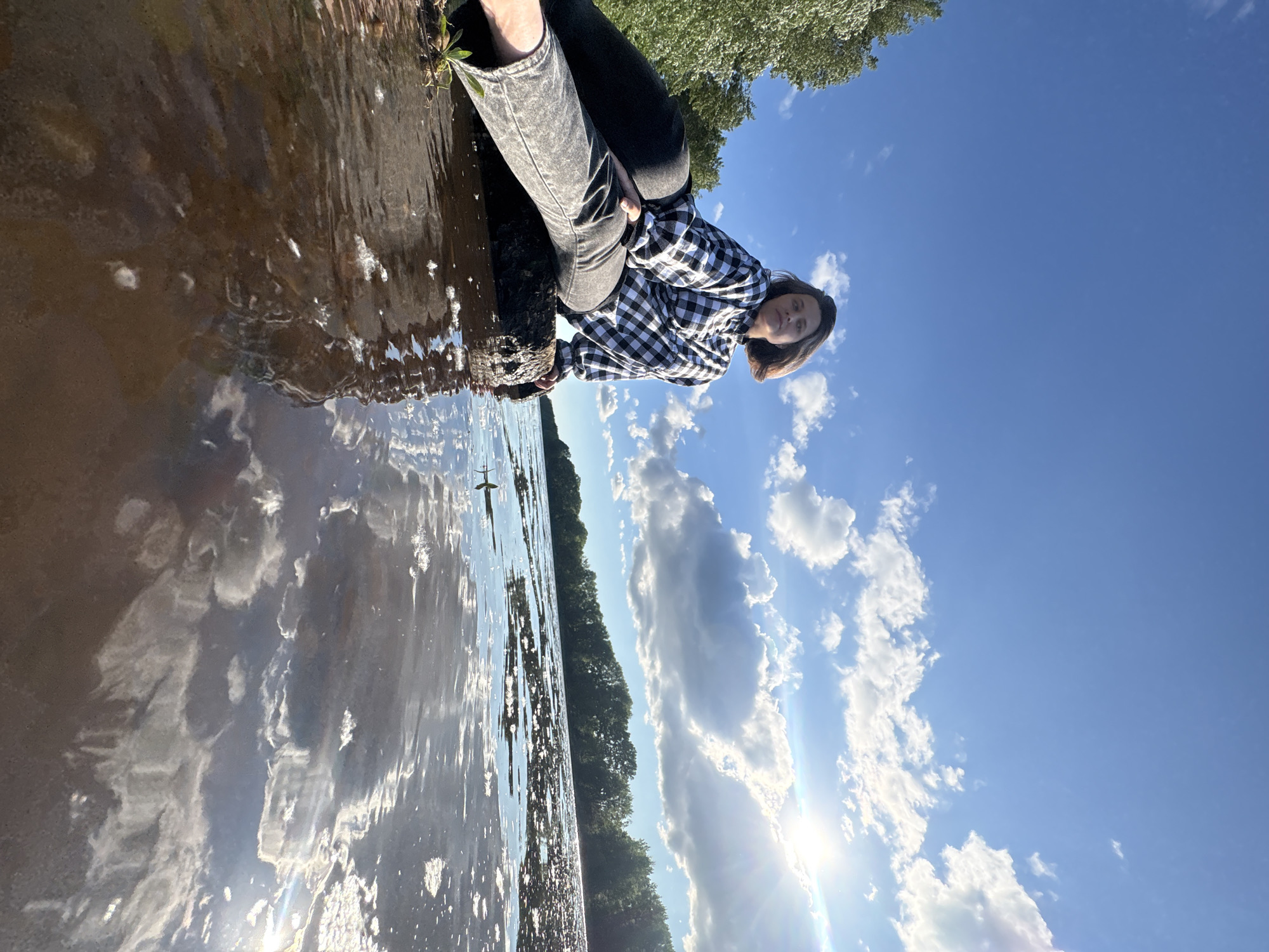A young woman with short brown hair is sitting on a large rock by the water. She is wearing a black and white checkered shirt and grey jeans. Her body is angled towards the right of the frame, and she is looking towards the camera. The water in front of her is brown and reflects the sky and clouds. The sky is blue with white, fluffy clouds. There are trees in the background on the far side of the water. The sun is shining brightly, creating a lens flare in the upper right corner of the image. The overall scene is peaceful and natural.