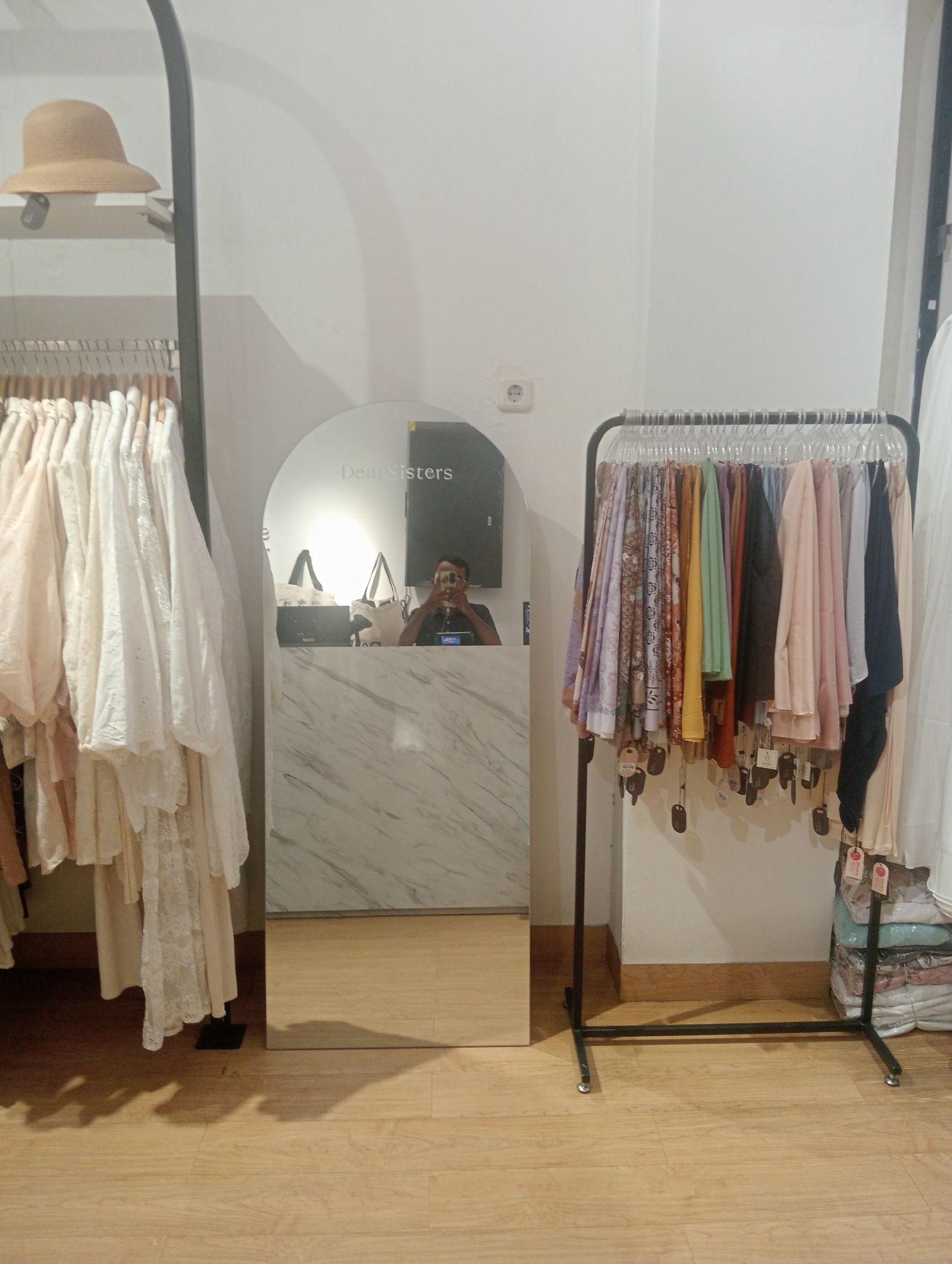 In a retail store, likely located in Larangan, Indonesia, a person is reflected in a large, arched mirror. The reflection shows the person holding a smartphone up to take a selfie. The mirror is positioned against a marble-patterned counter. To the left of the mirror, a clothing rack holds several white and cream-colored garments. Above this rack, a brown, wide-brimmed hat sits on a shelf. To the right of the mirror, another clothing rack displays an assortment of colorful scarves or hijabs, with price tags hanging from them. A stack of folded clothing is visible behind this rack. The floor is made of light-colored wood laminate. The walls are painted white. A white electrical outlet is visible on the wall to the left of the mirror. The overall impression is that of a boutique or clothing store display area.