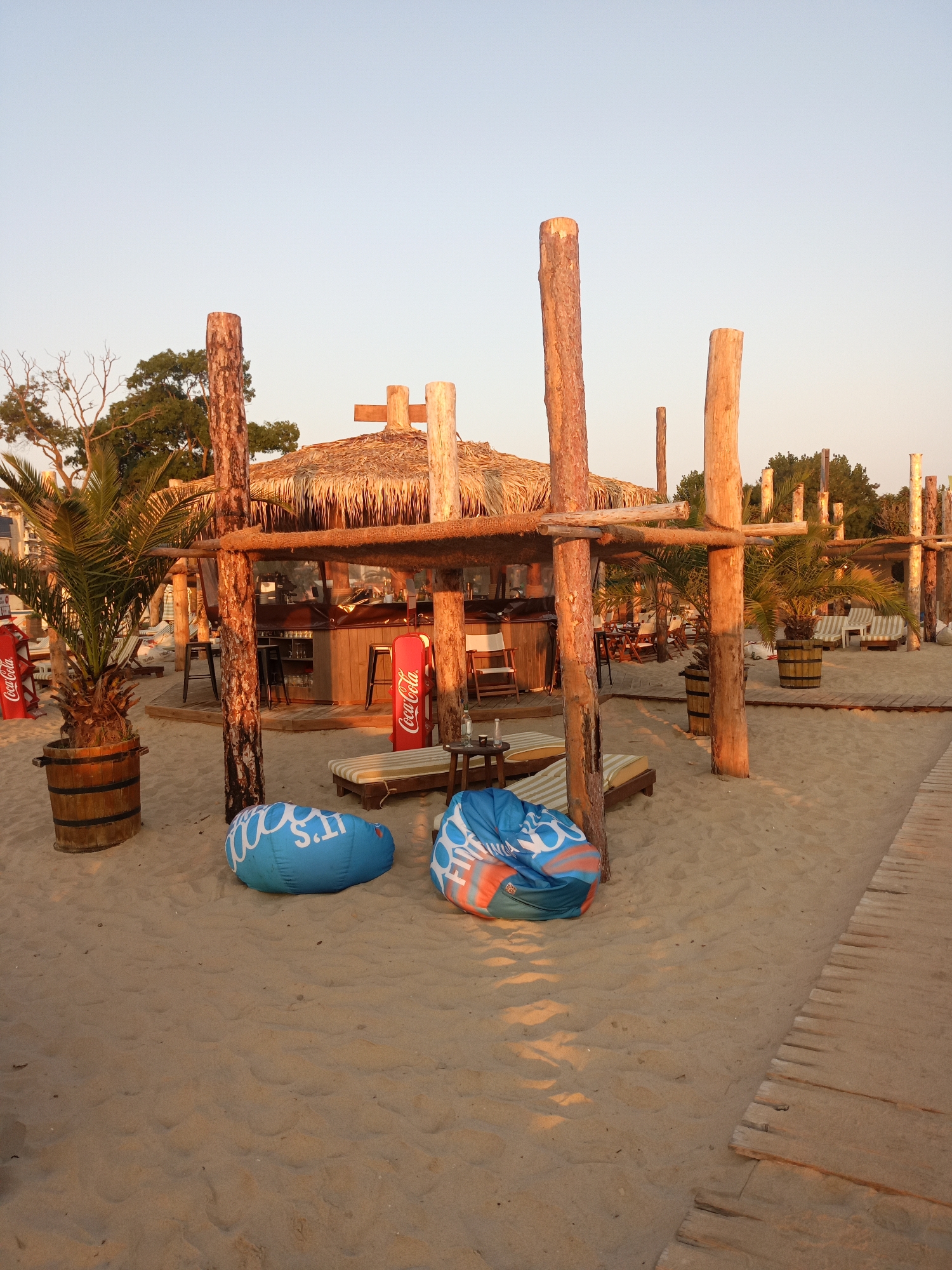 The image depicts a beach bar in Kranevo, Bulgaria, during a sunset. The scene is set on a sandy beach with a clear sky overhead. In the foreground, there are two large, inflatable beanbag chairs, one blue and white with the text "IT'S" visible, and another with blue, orange, and white patterns. A wooden boardwalk runs along the right side of the image, leading further down the beach.
Dominating the mid-ground is a rustic beach bar constructed with natural wood beams and a thatched roof. Palm trees are planted in wooden barrels next to the bar. Several lounge chairs with striped cushions are visible behind the beanbag chairs and near the bar, suggesting a seating area for patrons. Two red Coca-Cola advertising stands are placed on either side of the bar entrance. The warm, golden light of the sunset casts long shadows across the sand.
In the background, more trees and structures can be seen, indicating the presence of a resort or other facilities along the coastline. The overall atmosphere is relaxed and inviting, typical of a beachfront establishment in the late afternoon or early evening. There are no people visible in the image.
