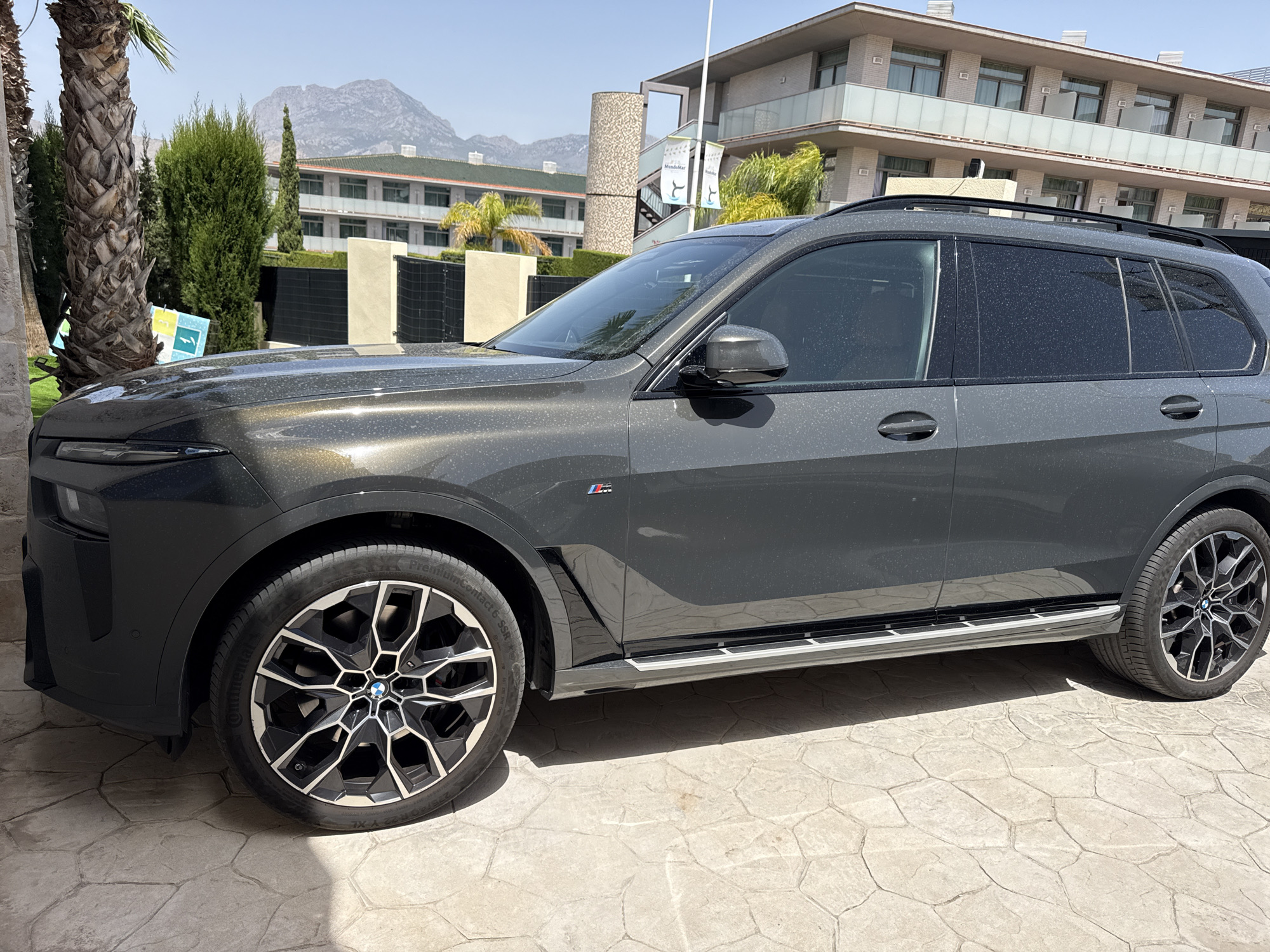 In an outdoor, sunlit environment, a dark grey BMW SUV is parked on a textured, light-colored paved surface. The car's exterior is dusty, with numerous small white specks visible, particularly on the dark paintwork. The vehicle features distinctive black alloy wheels with a complex, multi-spoke design and a silver trim along the lower body panels. A BMW logo is present on the wheels.
Behind the car, a multi-story building with a modern architectural style is visible. It has a light-colored facade, multiple balconies with glass railings, and windows. Green trees and palm trees are scattered around the building and in the foreground, with one large palm tree trunk prominently on the left side of the image. In the background, a large, rocky mountain range under a clear blue sky completes the scene. Banners with text and a wave-like graphic are hanging from a pole near the building. The overall impression is one of a resort or hotel setting.