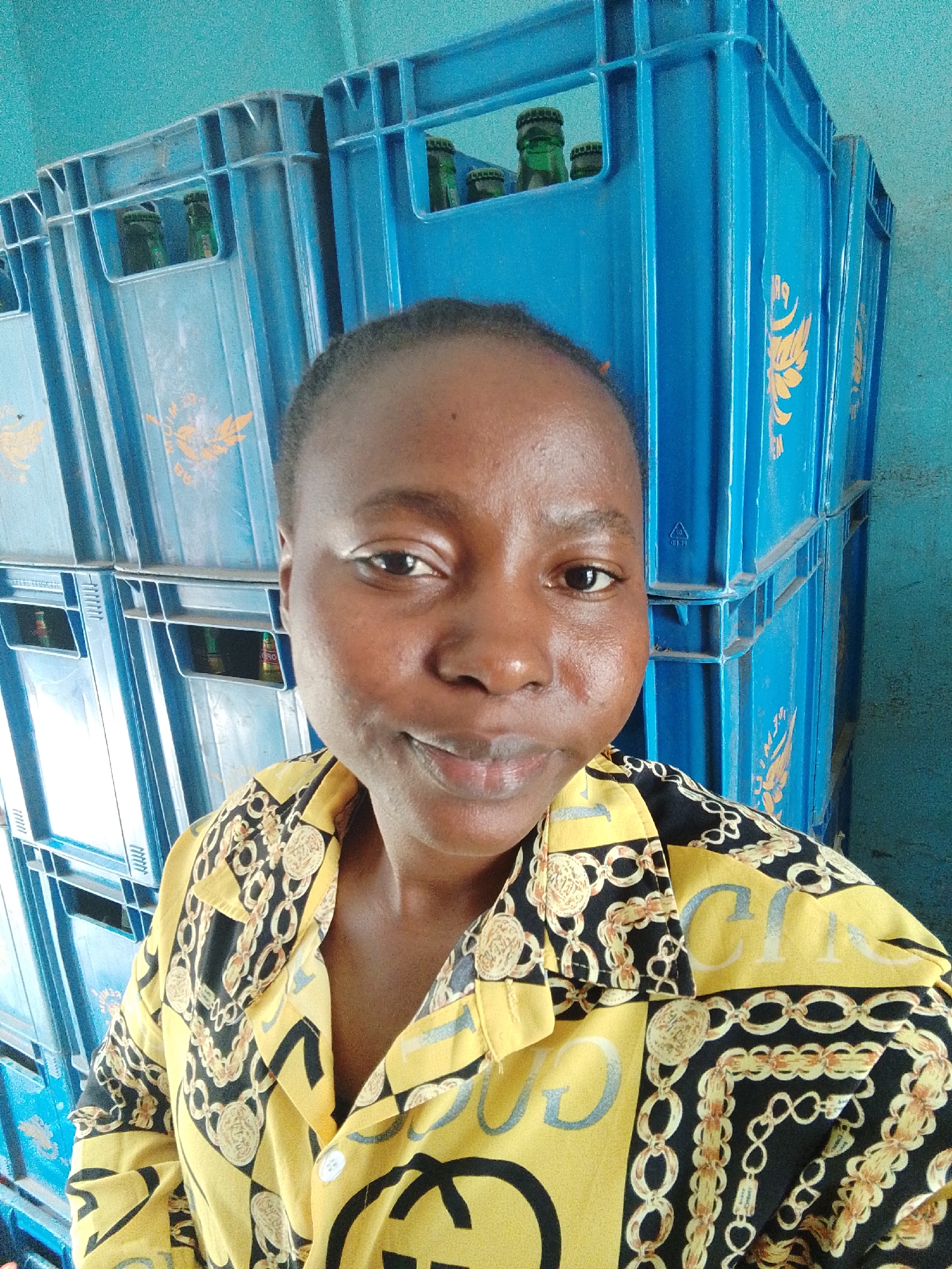A woman with short, dark hair and dark eyes is centered in a close-up, eye-level selfie. She looks directly at the camera with a gentle smile. She is wearing a yellow and black collared shirt with a pattern featuring gold chains, coins, and the letters "GVC" or "GUCCI" in yellow. The background shows tall stacks of blue plastic crates, some of which contain green glass bottles, likely beverages. The crates have a yellow or gold eagle/bird emblem and some indistinct text, including a recycling symbol on one crate. The setting appears to be an indoor space, possibly a storage area or a shop, with a blue wall behind the crates. The lighting is bright and even, suggesting it is daytime.