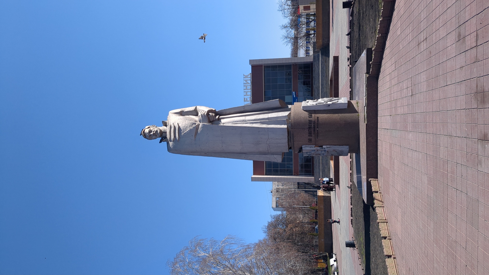 Here is a detailed description of the image:
The image is of the statue of G.D. Gay, a Soviet writer, located in Ulyanovsk, Russia. The statue is a full-body depiction of the writer, standing and draped in a long coat. The statue is made of a light-colored material, possibly stone or concrete. It stands on a cylindrical pedestal, which also contains text and other sculptural elements.
Behind the statue is a building with a glass facade. The building has the Russian word "ЛЕННИК" on top. The sky is clear and blue, with a bird flying in the distance.
In the foreground, there is a paved area with red bricks. The area seems to be a public square or park. There are some trees in the background, but they appear bare, suggesting it might be winter or early spring. There are also a few people walking around in the background.
