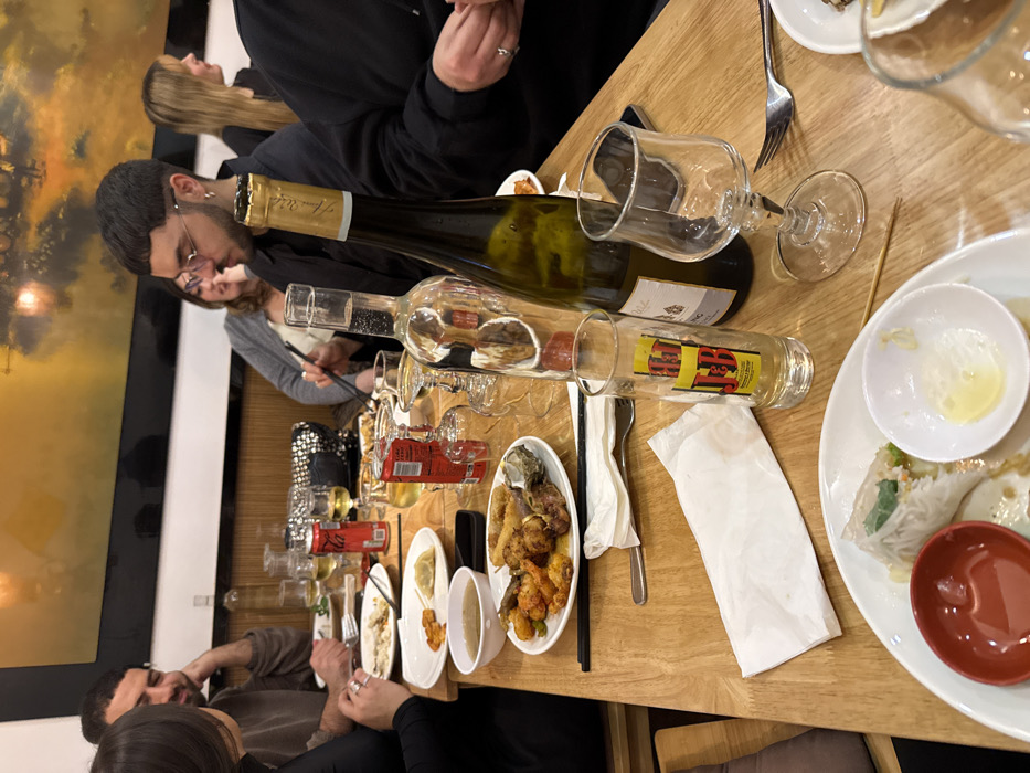 A large wooden table covered with food and drinks sits in a restaurant. There are two wine bottles, several glasses, and various dishes, including spring rolls, dumplings, and fried food. Several people are gathered around the table, eating and talking. The scene is lively and social, with a sense of enjoyment and camaraderie. A painting is on the wall.