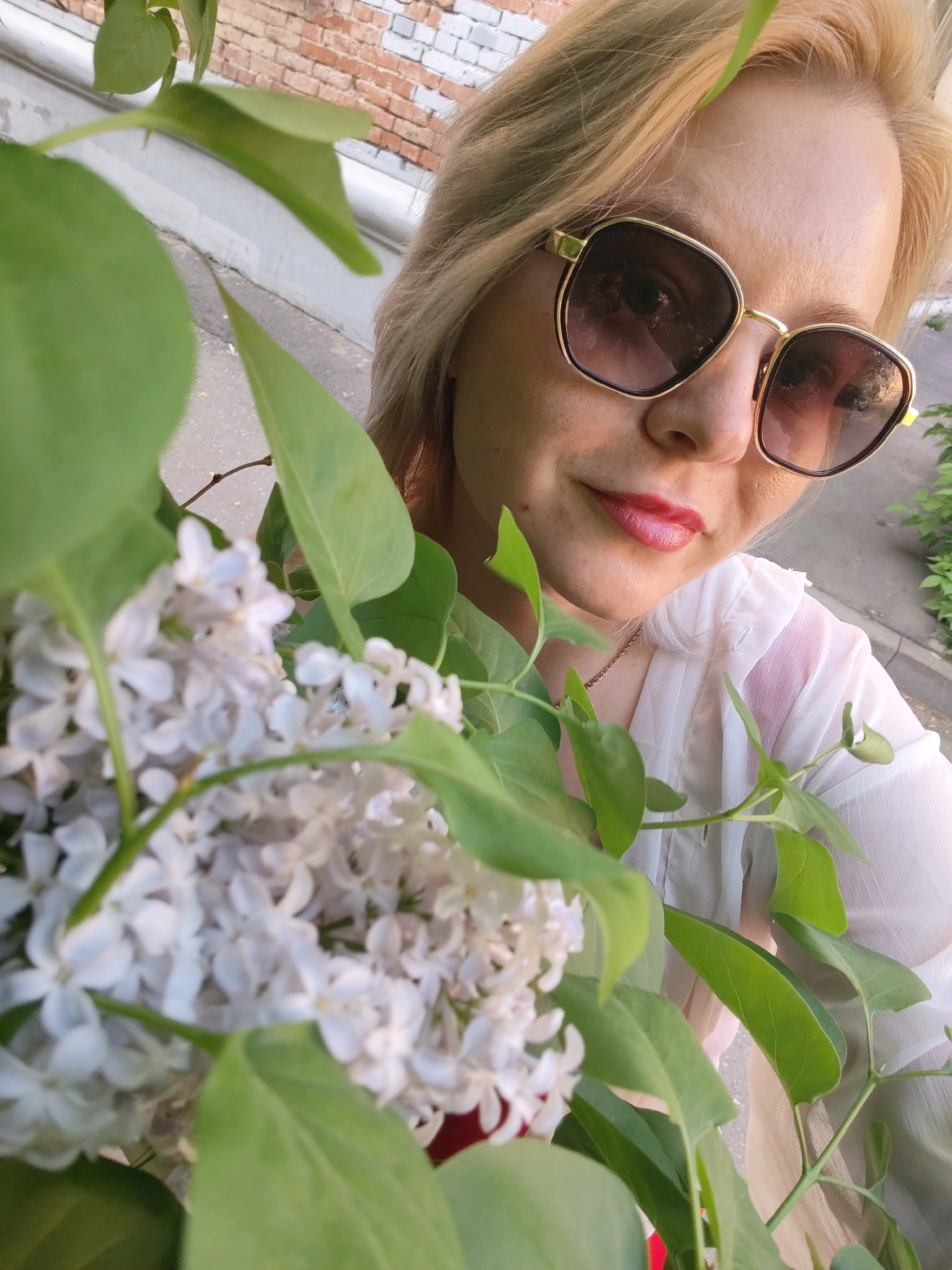 A fair-skinned woman with short blonde hair is holding a bouquet of white lilacs in front of her. She is wearing sunglasses with dark lenses and a thin gold frame and red lipstick. Her top appears to be a translucent white fabric. Behind her is a section of a brick wall, possibly belonging to a building. There is a narrow strip of asphalt visible in the background, indicating she may be standing near a street or sidewalk. Green foliage is also seen to the right. Based on the available information, the exact location in Izhevsk, Russia cannot be precisely determined.