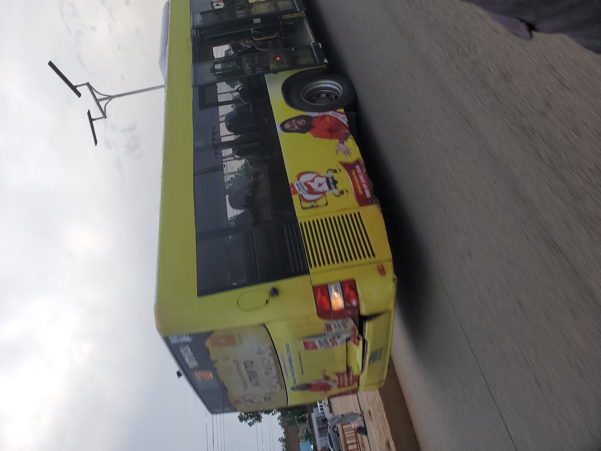 A yellow bus is captured in motion on a road, likely in Bukuru, Nigeria, under an overcast sky. The bus, which appears to be a commercial passenger vehicle, displays advertisements on its sides. On the rear of the bus, there is visible damage to the bumper, suggesting a recent impact. The bus has tinted windows and its doors are open. In the background, there are glimpses of other vehicles, including a white van and a silver car, and some buildings and vegetation. A street light with two solar panels is visible above the bus. The road surface is asphalt.